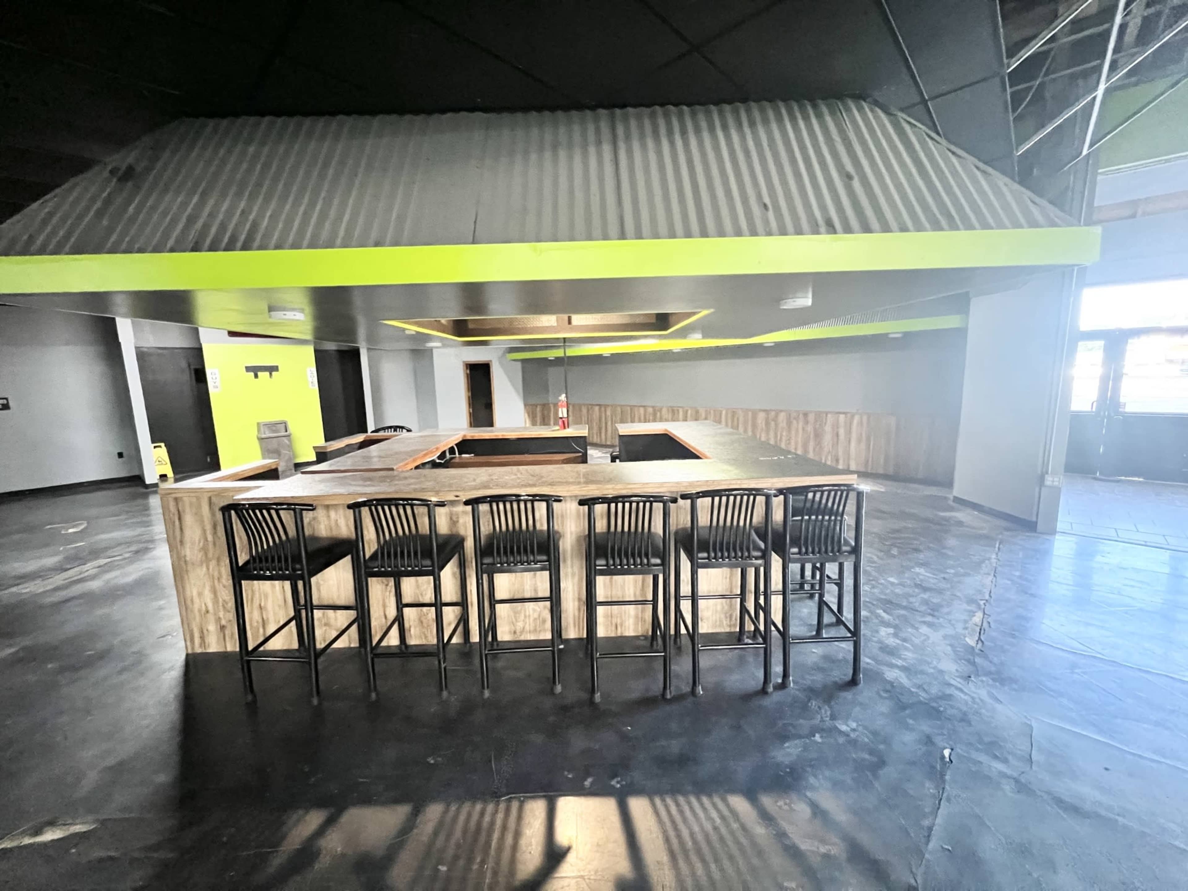 The image shows a spacious bar area with a U-shaped counter and several black stools lined up in front of it, situated in a room with a high ceiling and industrial-style design.