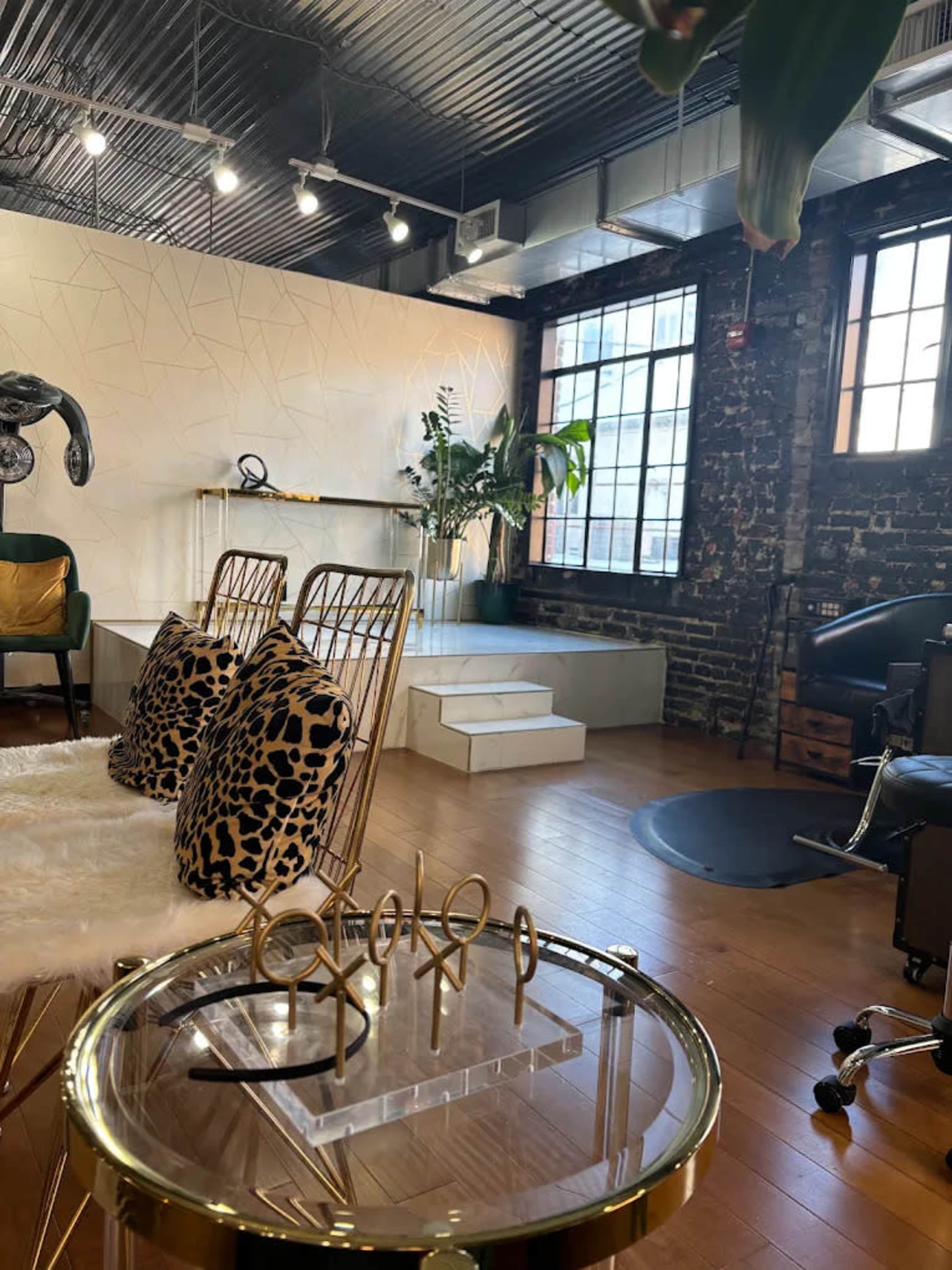 The image shows a stylish interior of a modern salon or studio featuring a combination of brick walls, large windows, and decorative furniture, including a glass table and gold-accented seating.