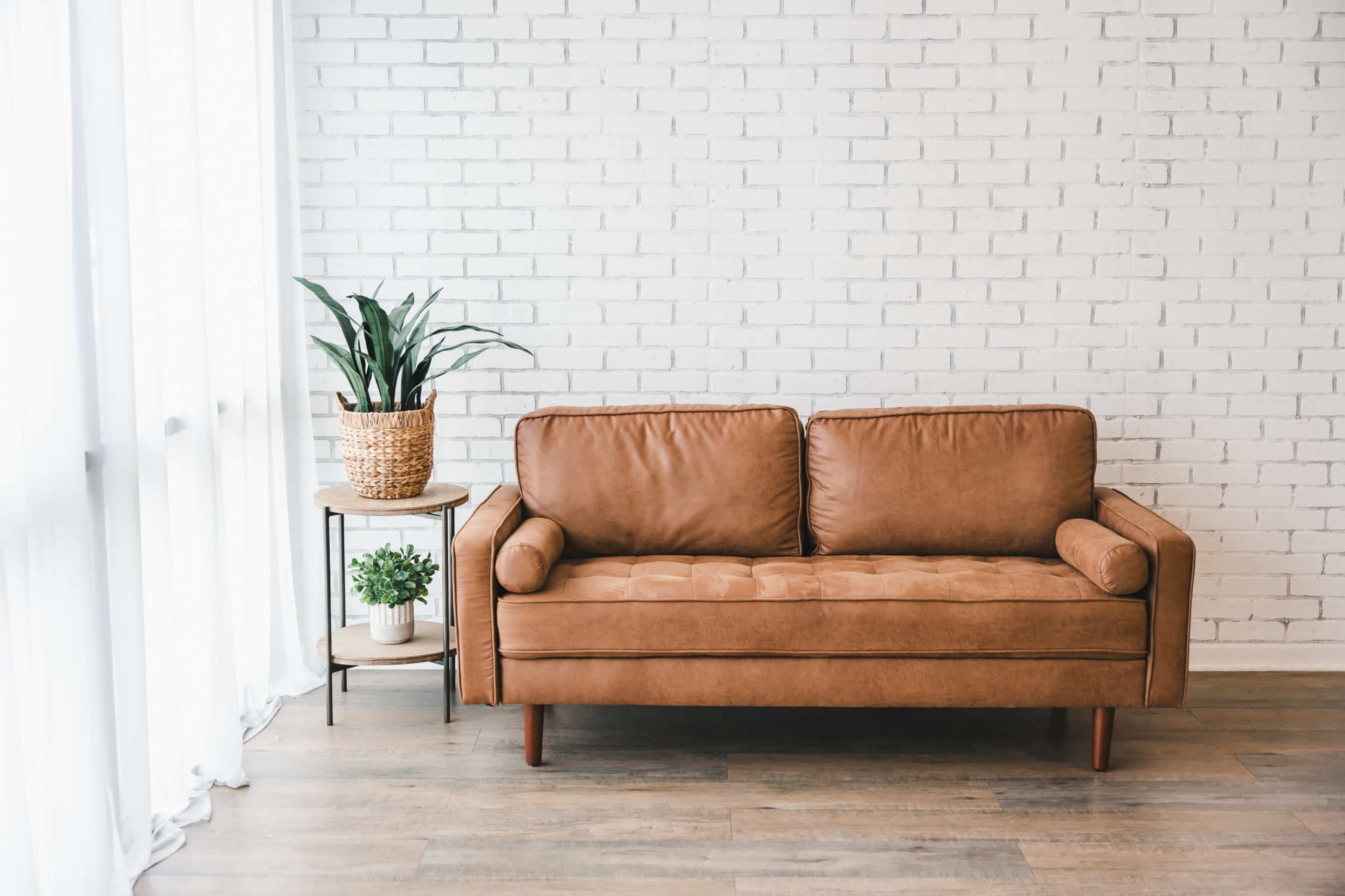 A brown leather couch sits against a white brick wall next to a potted plant on a small table.