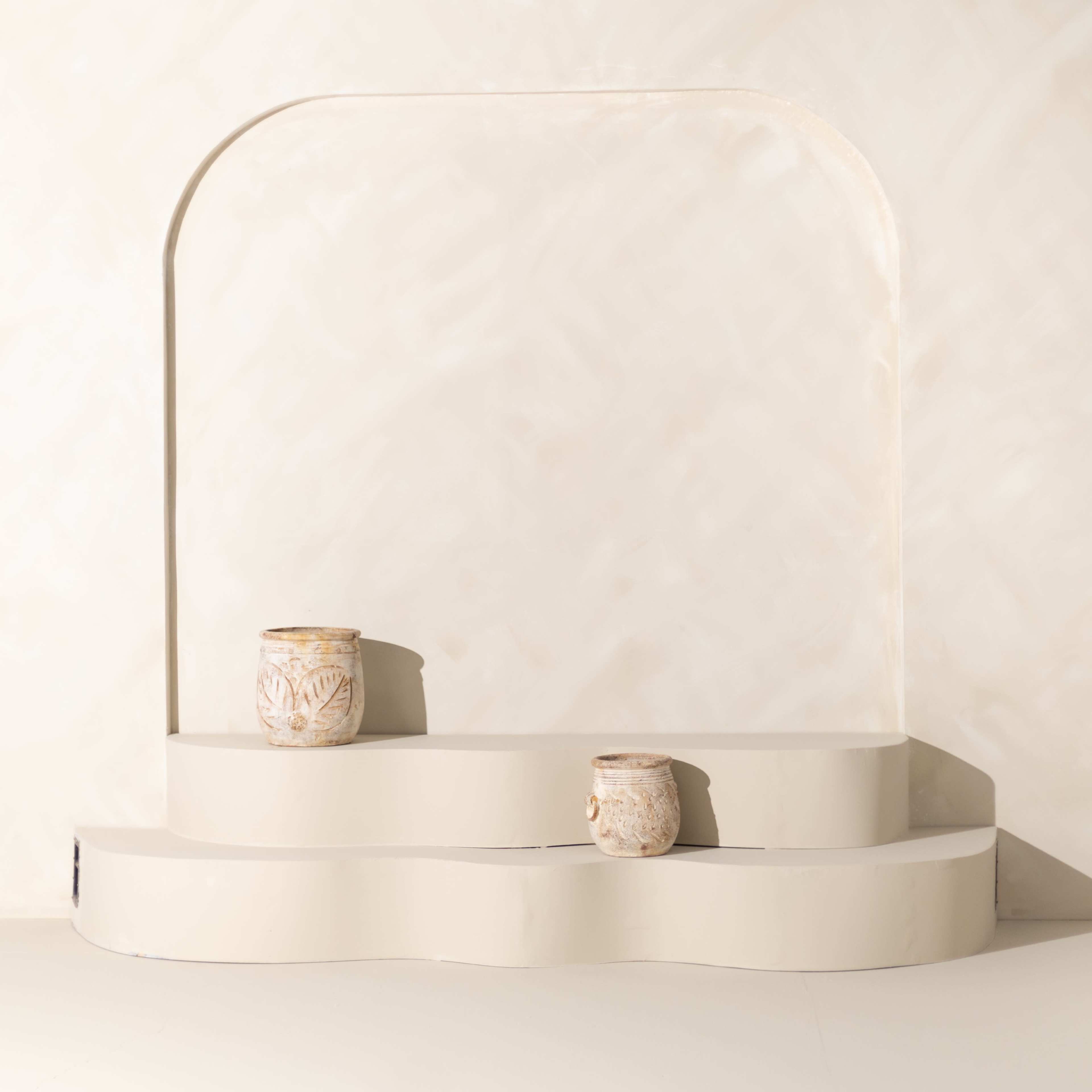 The image shows a minimalist display featuring two decorative pots on a tiered, curved platform against a light-colored wall.