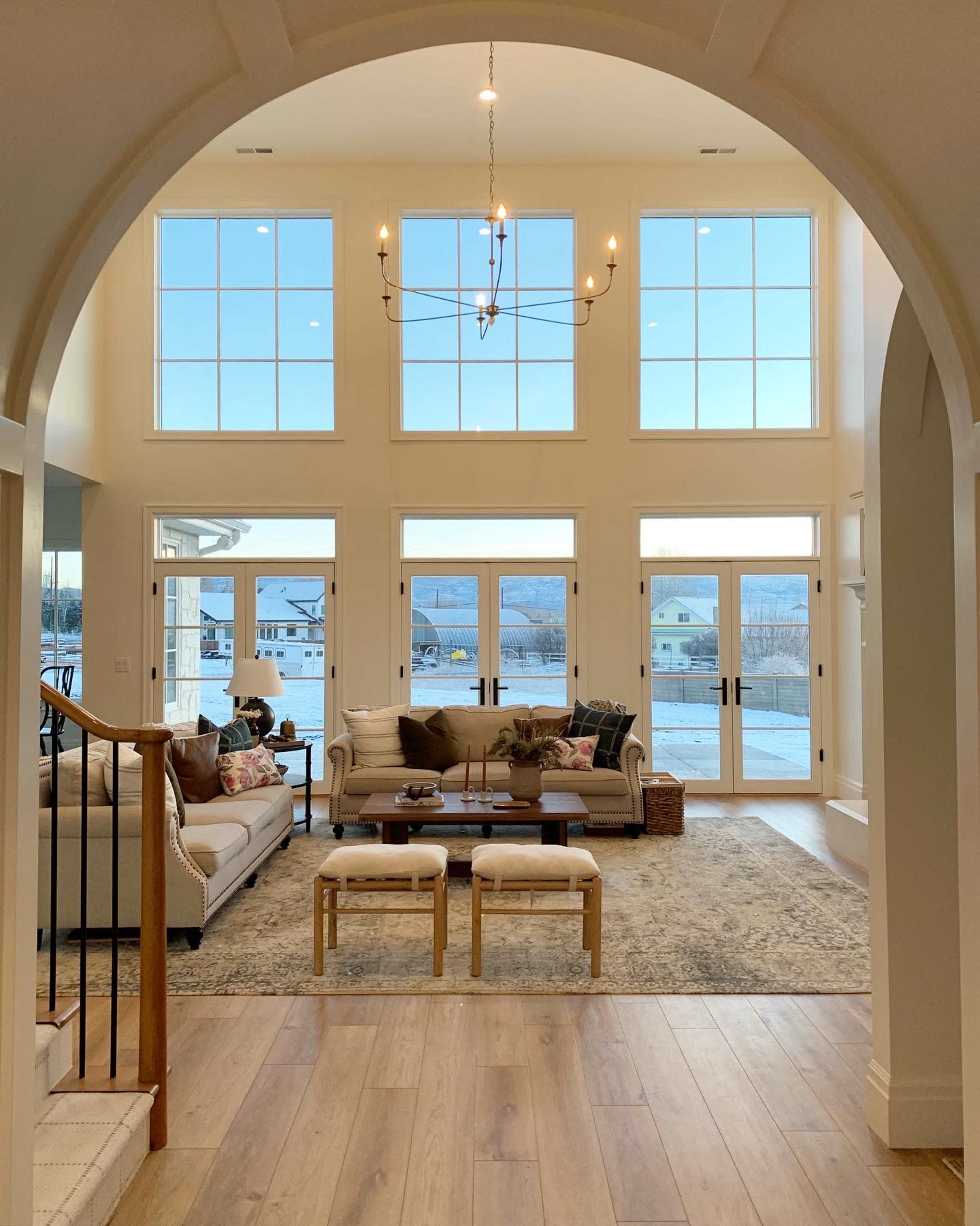 A spacious living room features large windows that allow natural light to fill the area, with a seating arrangement centered around a coffee table and a view of the outdoors.