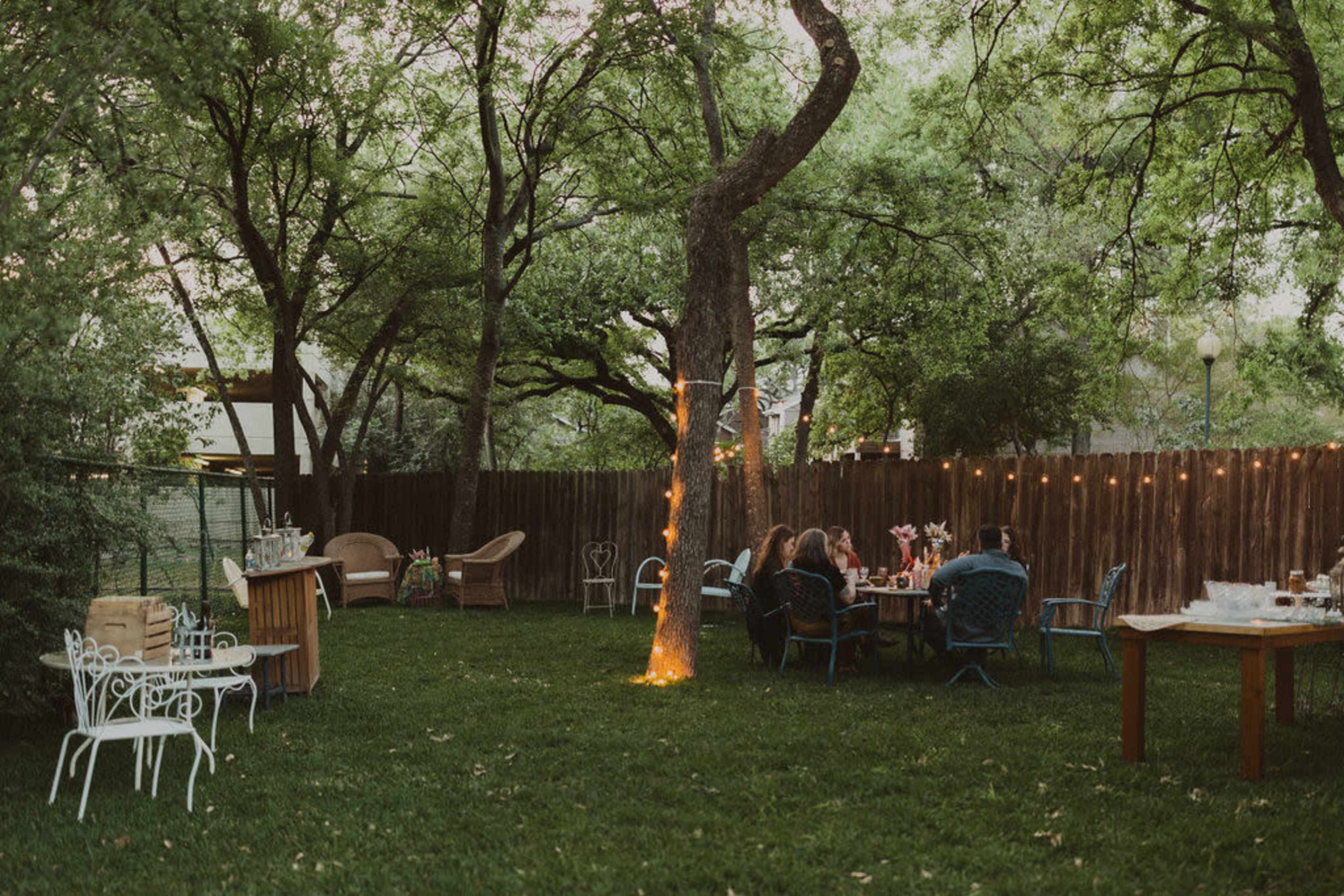 A serene outdoor gathering is set up in a lush backyard with various seating arrangements, a table adorned with flowers, and string lights illuminating the area.