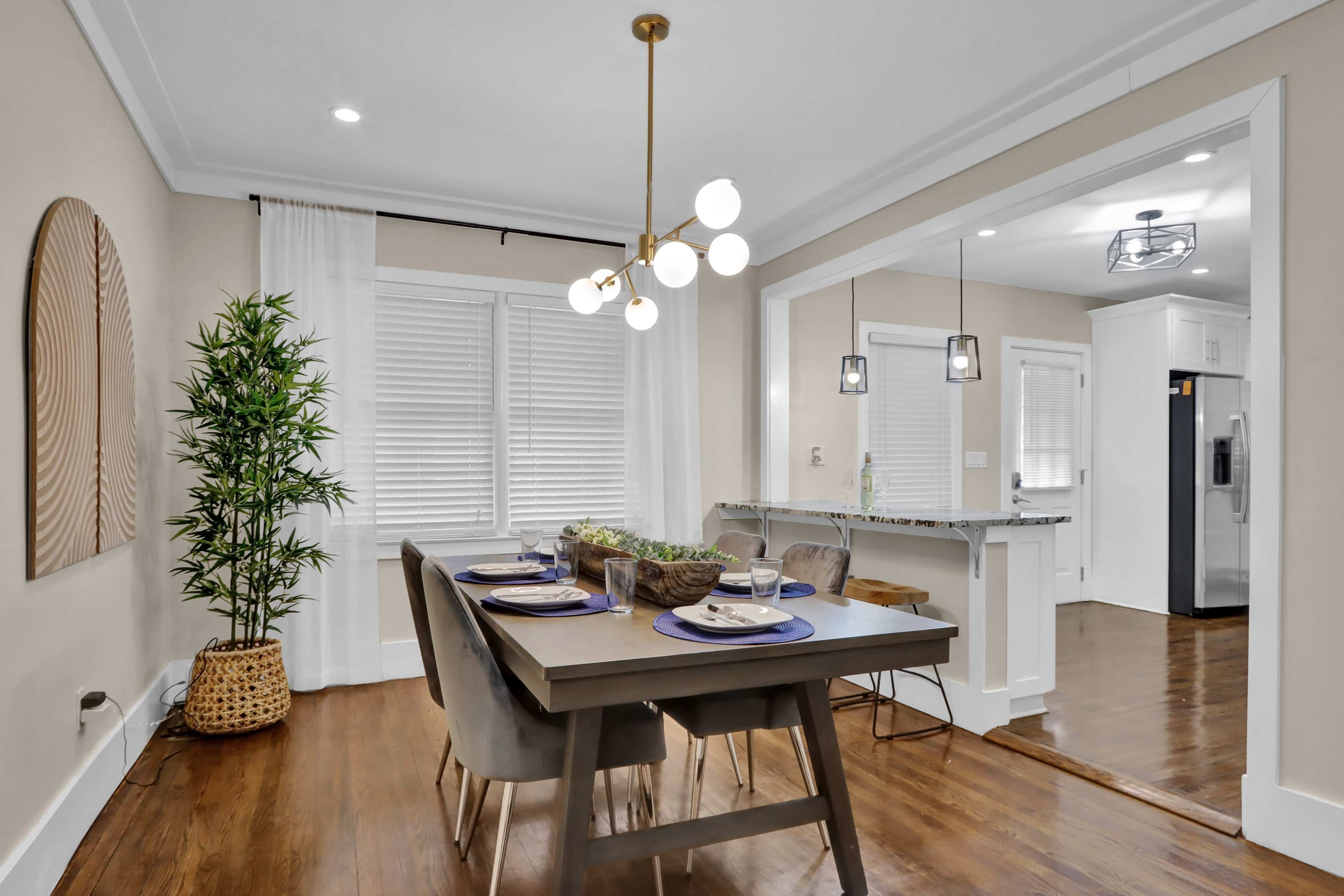 A modern dining area features a wooden table set for six, surrounded by chairs, with a large window and a kitchen bar in the background.