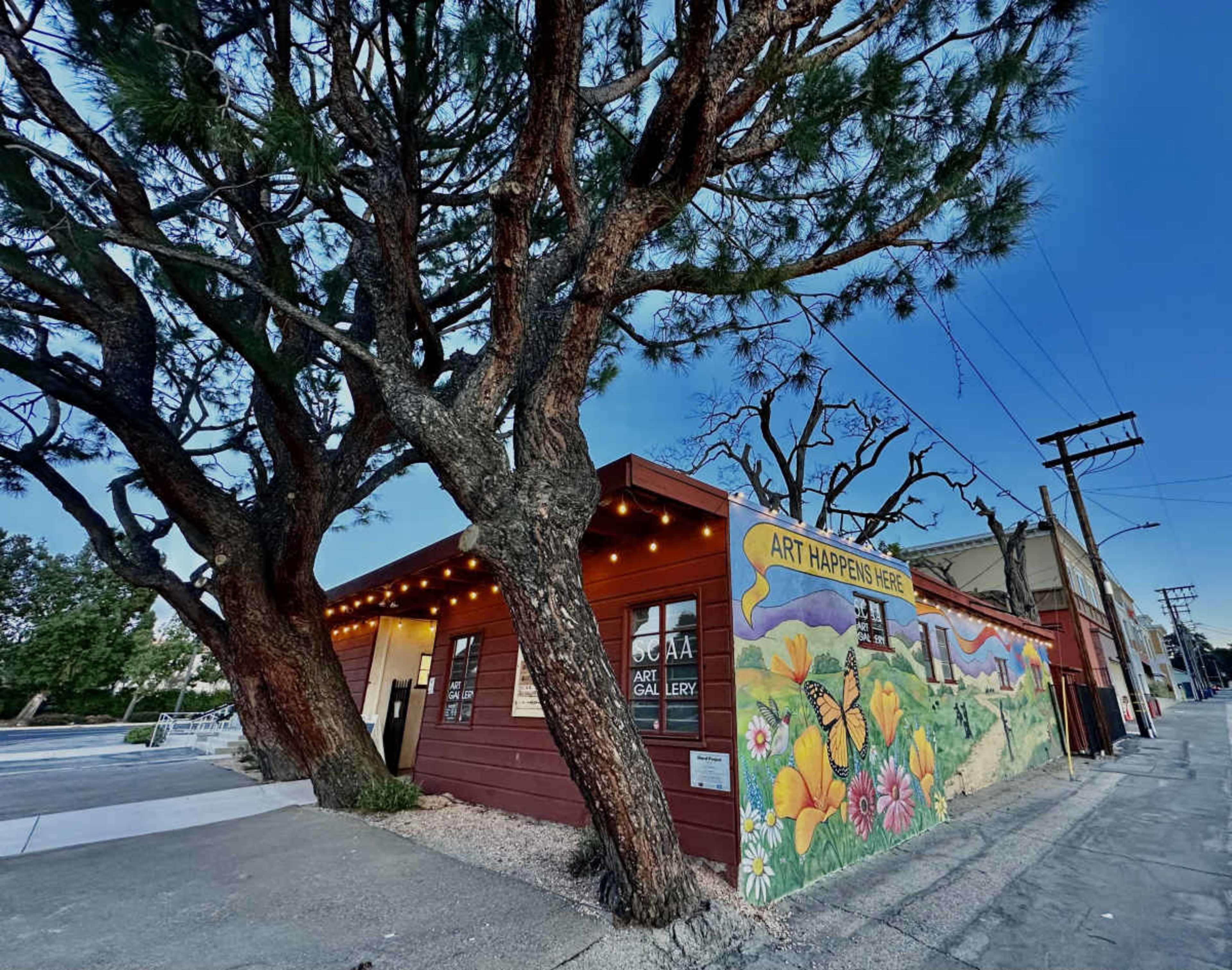 A modern art gallery with a colorful mural on its side is situated next to large trees and a clear sky.