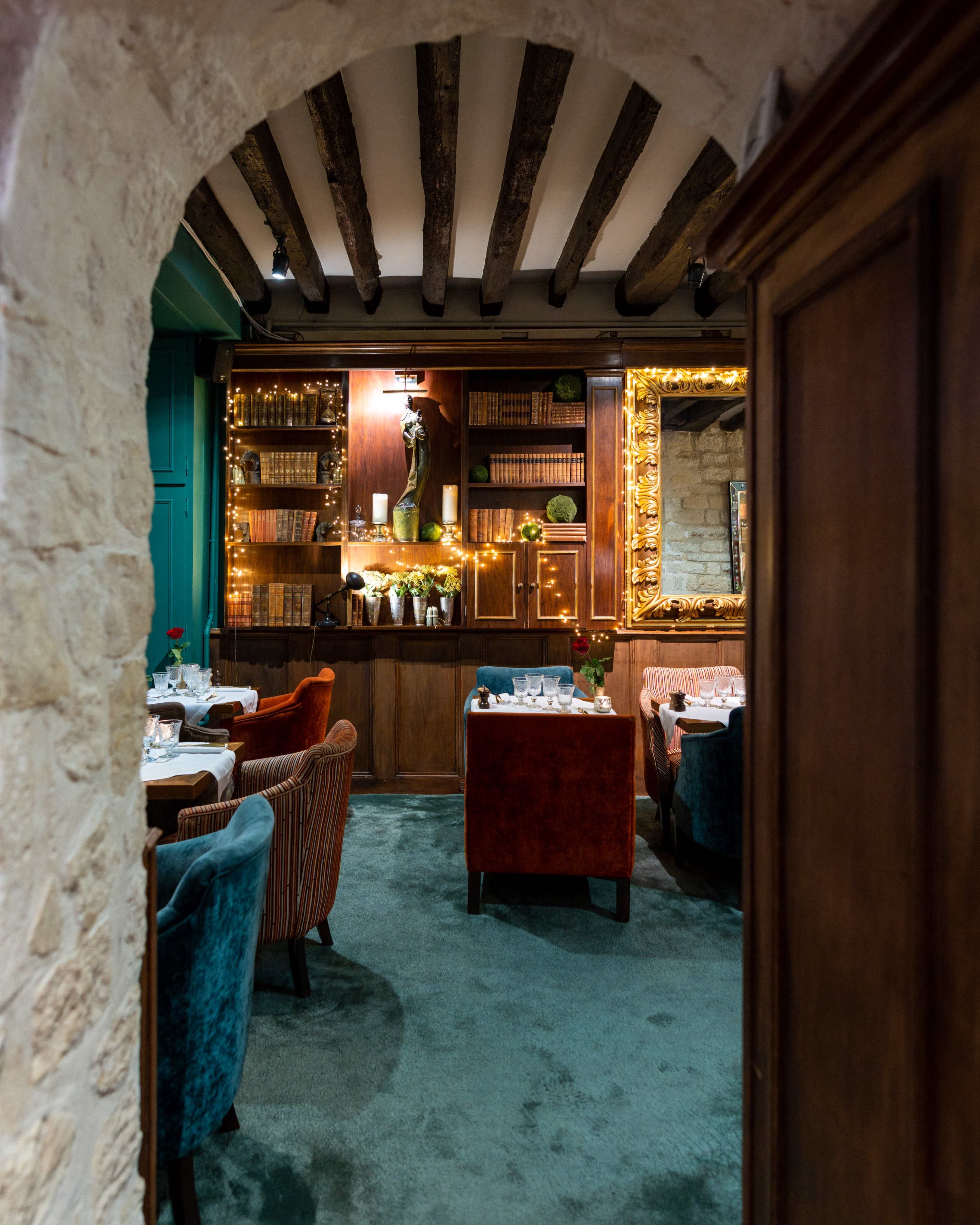 The image shows a cozy restaurant interior with wooden beams, shelves lined with books, and plush seating arranged around tables.