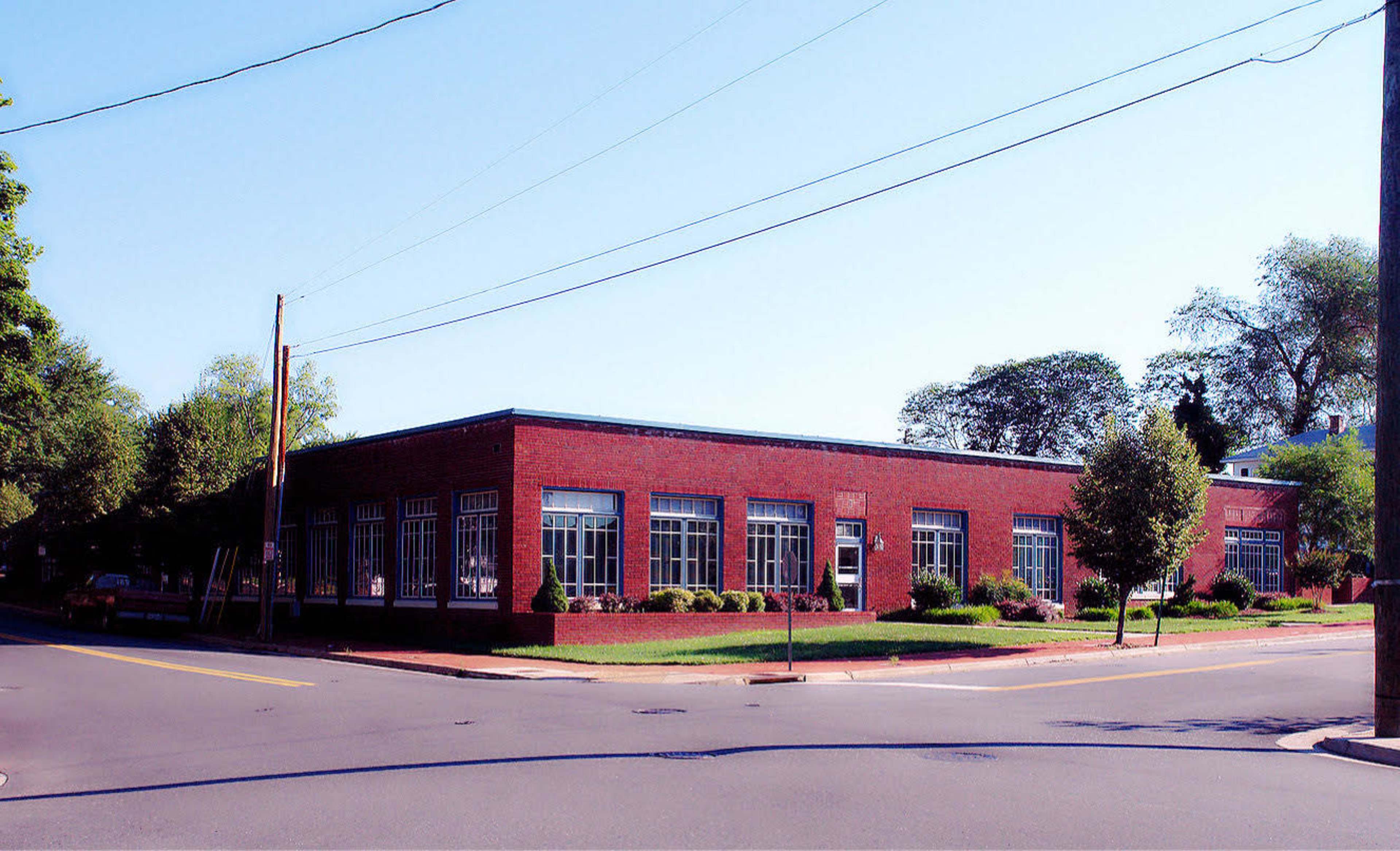 A single-story red brick building with multiple large windows is situated on the corner of a street, surrounded by neatly maintained landscaping.