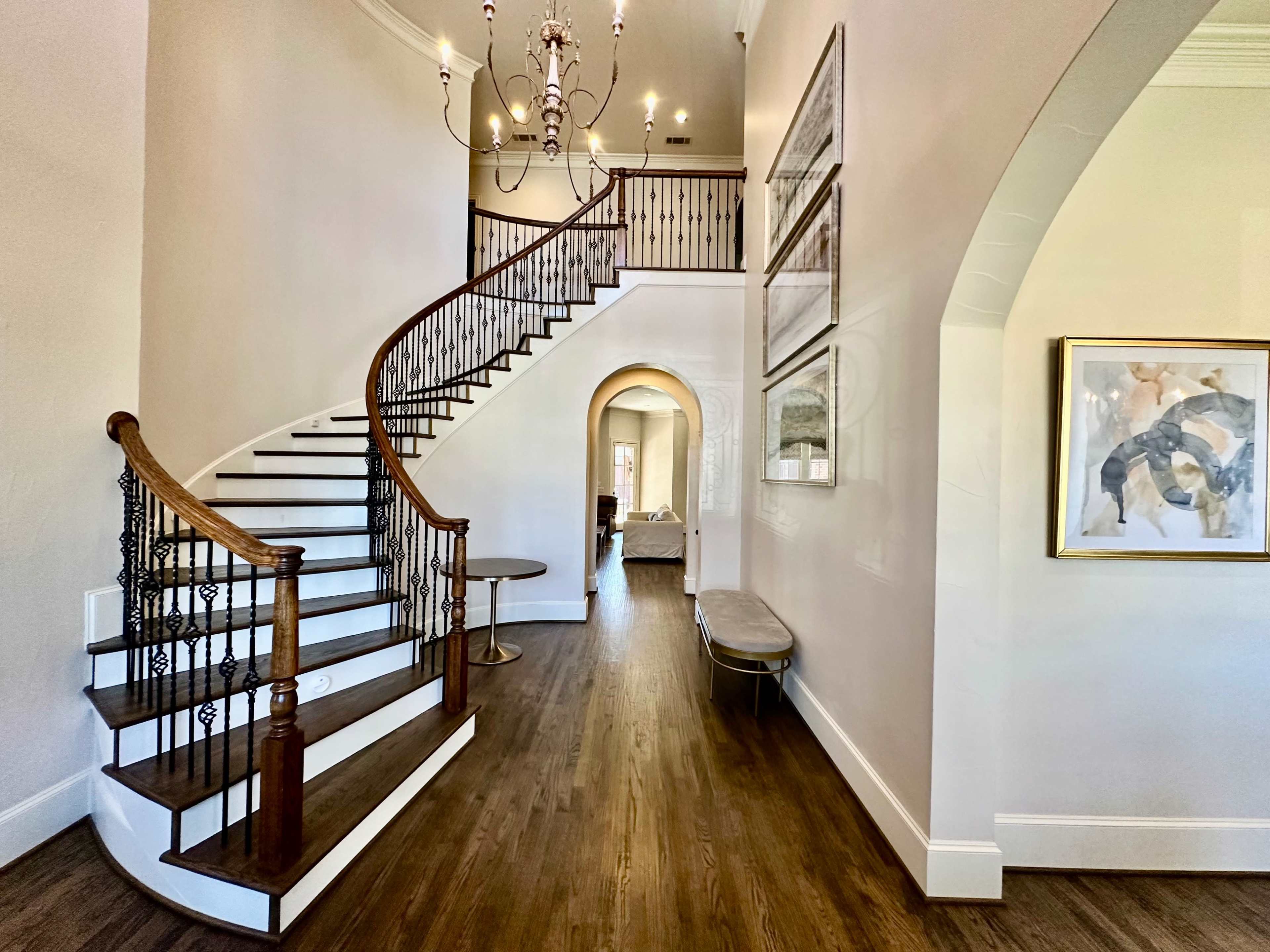 The image shows a well-lit, elegant foyer with a curved staircase, wooden flooring, and decorative wall art.
