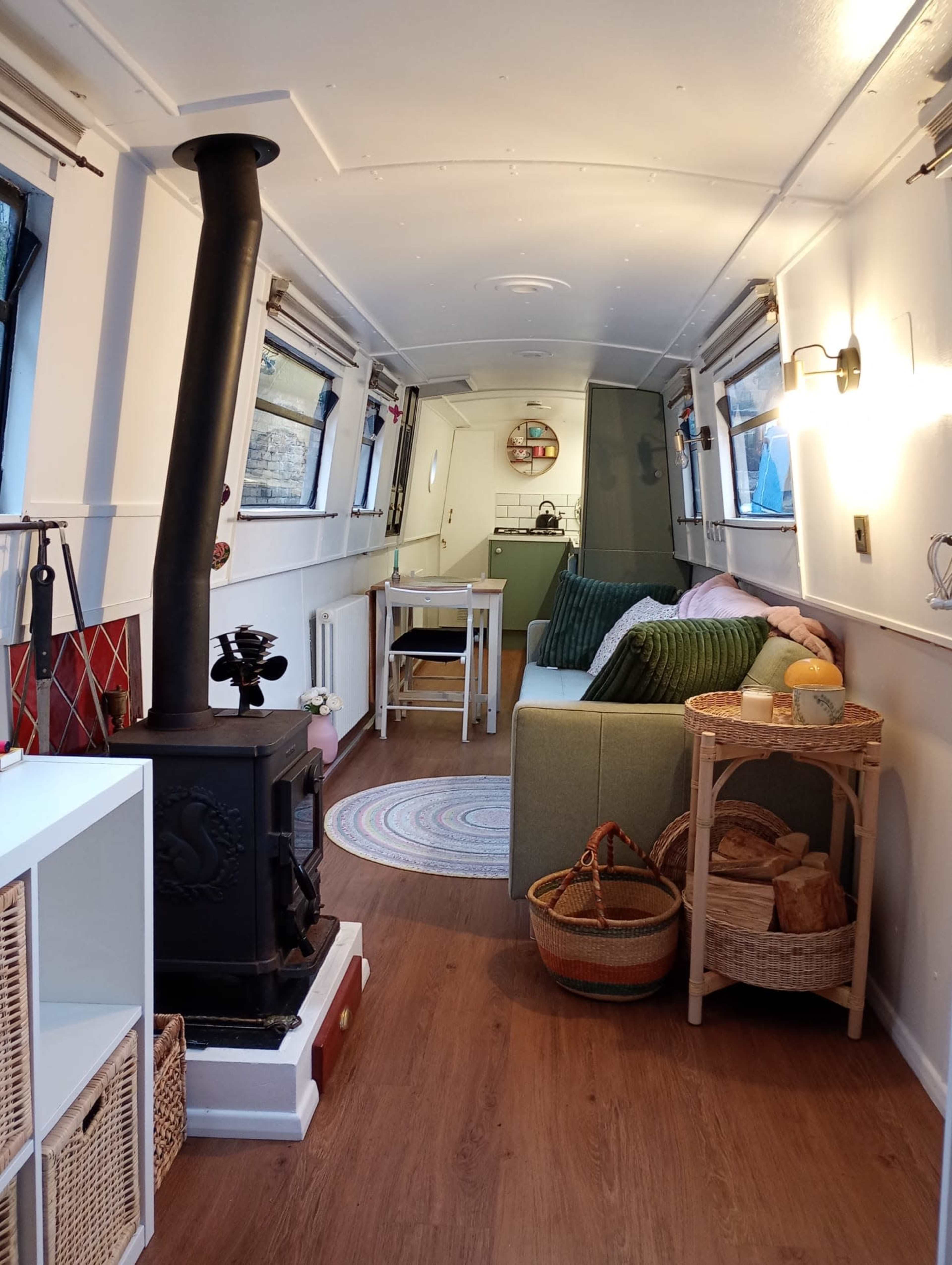 The interior of a narrowboat features a cozy seating area, a wood-burning stove, a small dining table, and decorative baskets.