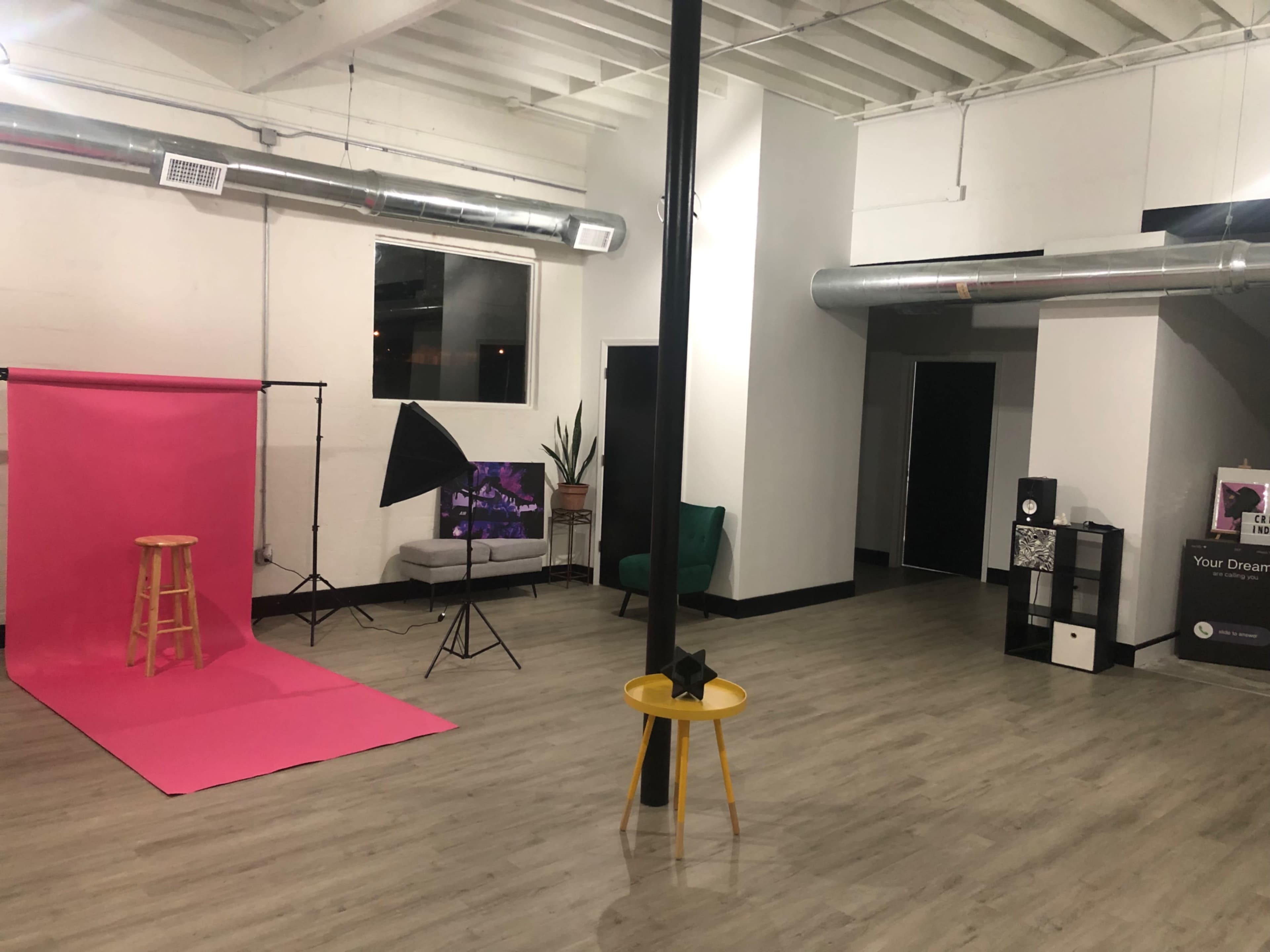 A spacious studio with a pink backdrop, a stool, a photography light, and a green armchair, along with a small black and white shelf in the corner.