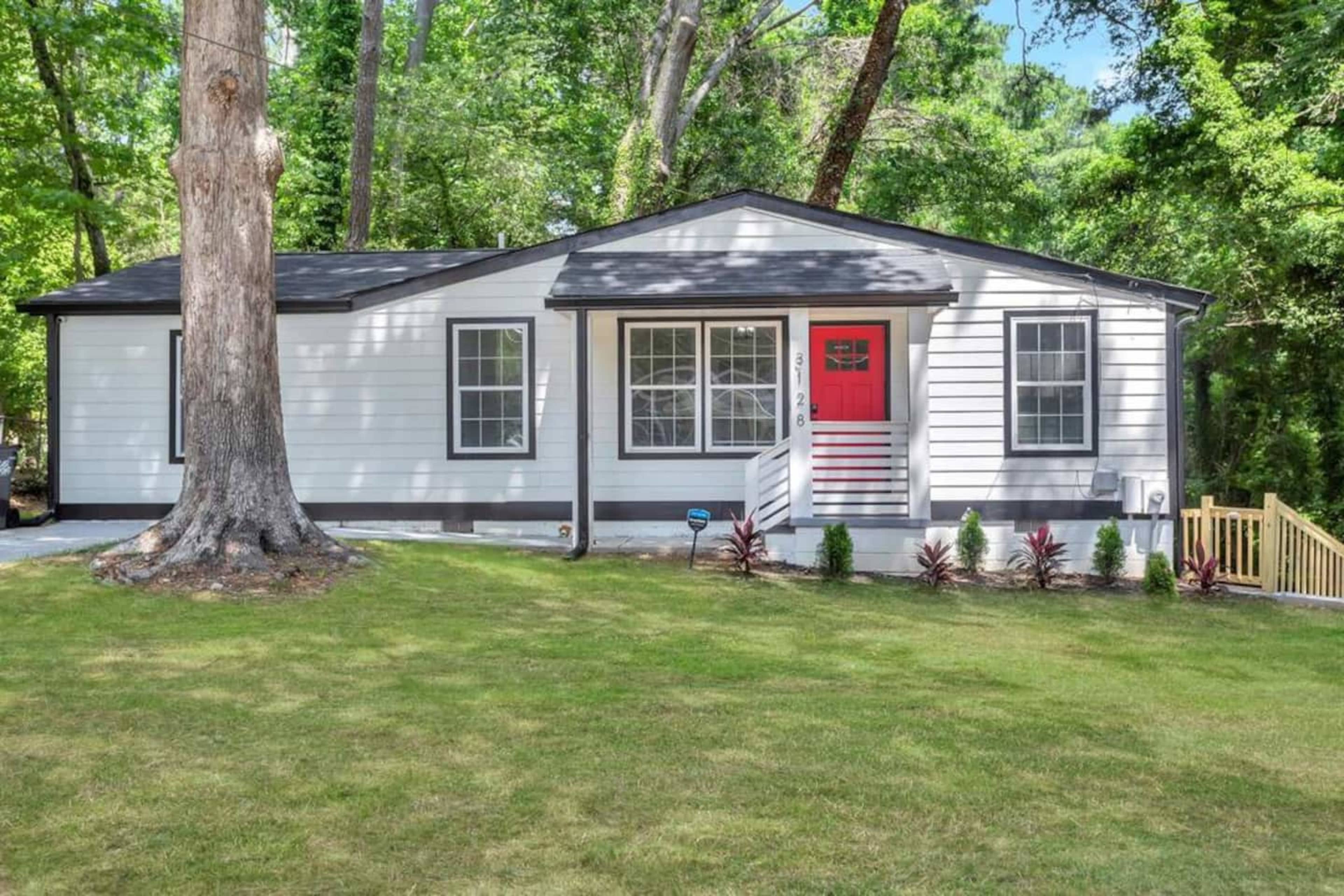 A single-story house with a gray and white exterior, red front door, and a lawn surrounded by trees.