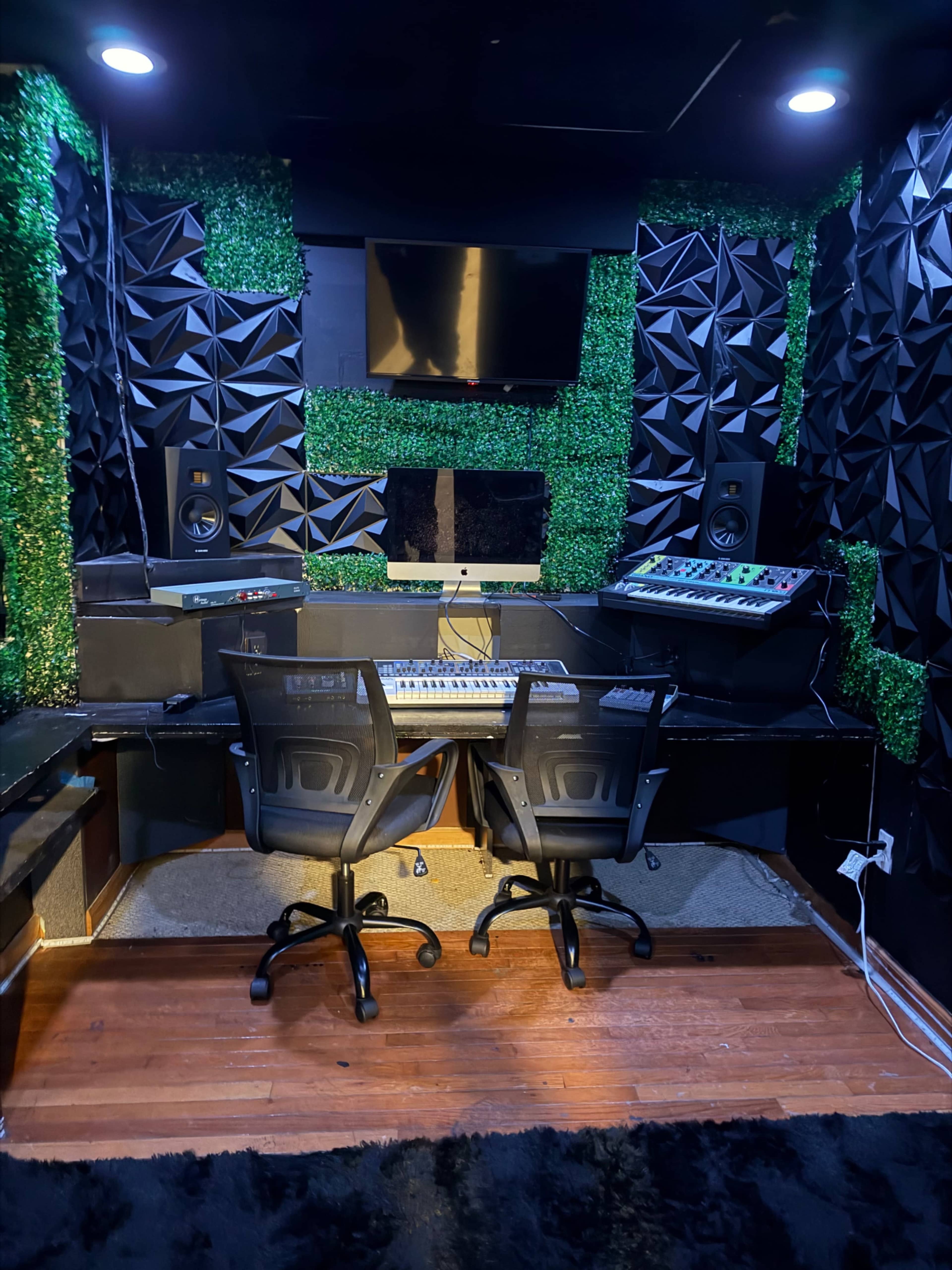 The image shows a music studio with two chairs facing a keyboard and various electronic equipment, surrounded by green textured wall panels.