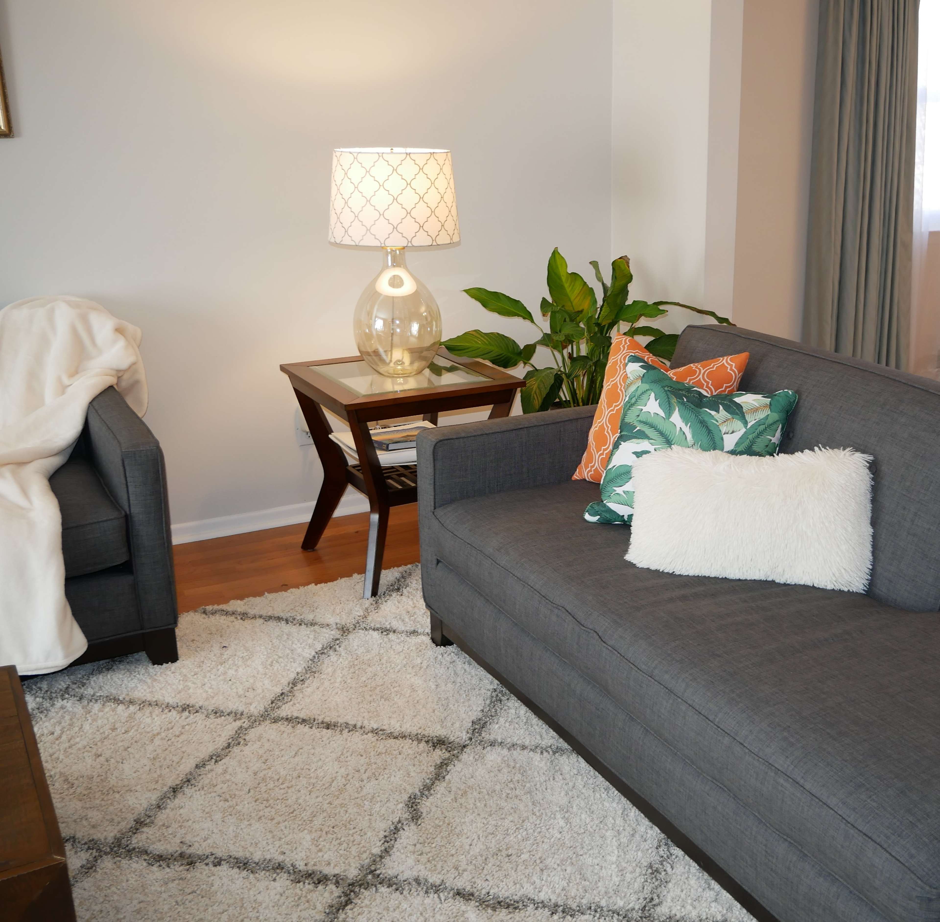 A modern living room with a gray sofa, a cozy armchair, a decorative table lamp, and a potted plant beside a small accent table on a light-colored rug.