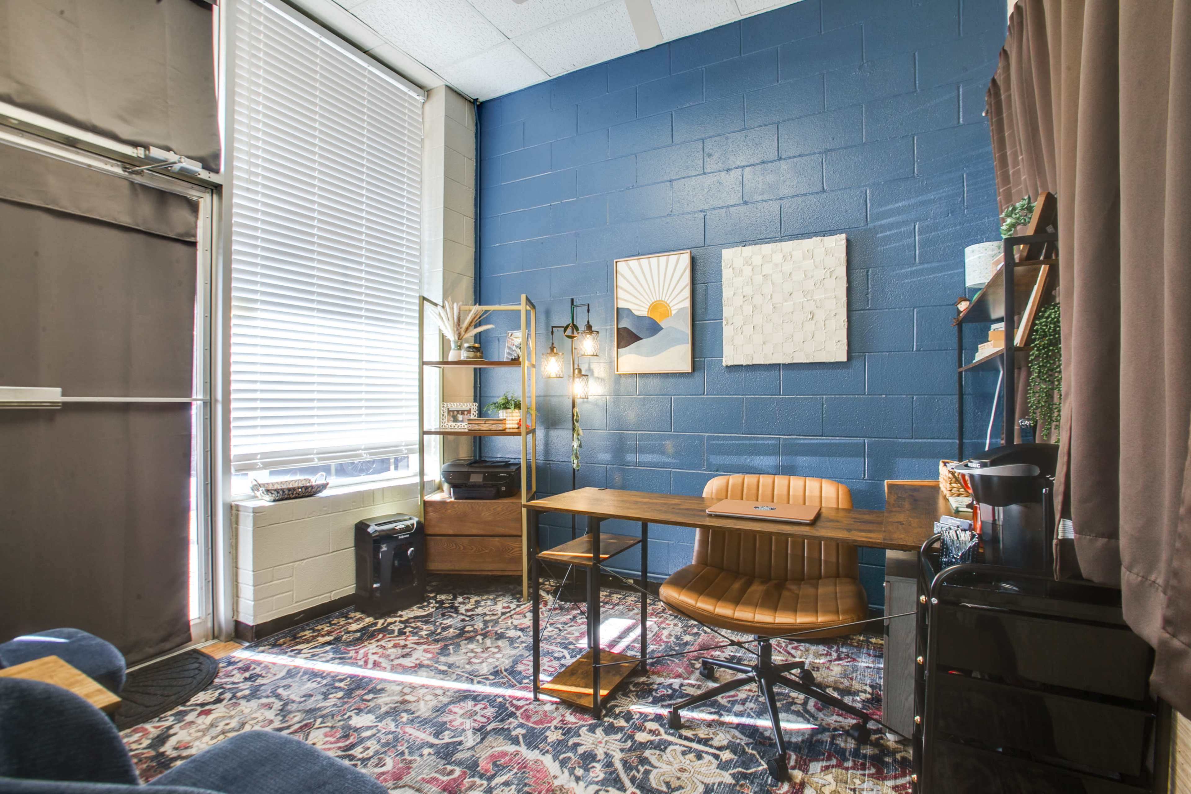 The image shows a small office space featuring a wooden desk with a chair, a bookshelf, and artwork on the wall, illuminated by natural light from a window.