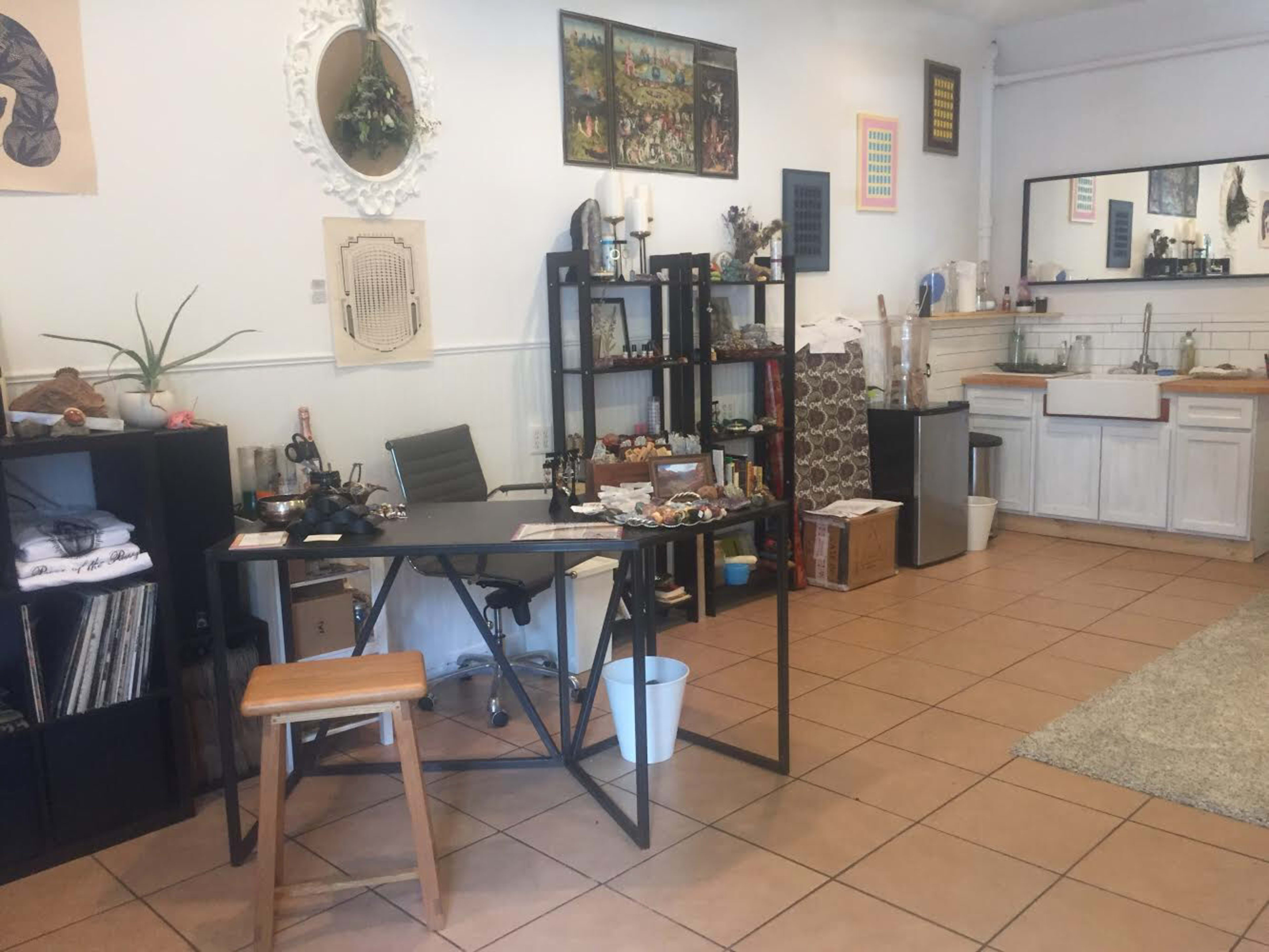 An interior space featuring a desk, a stool, shelving units filled with items, and a sink area along with tiled flooring.