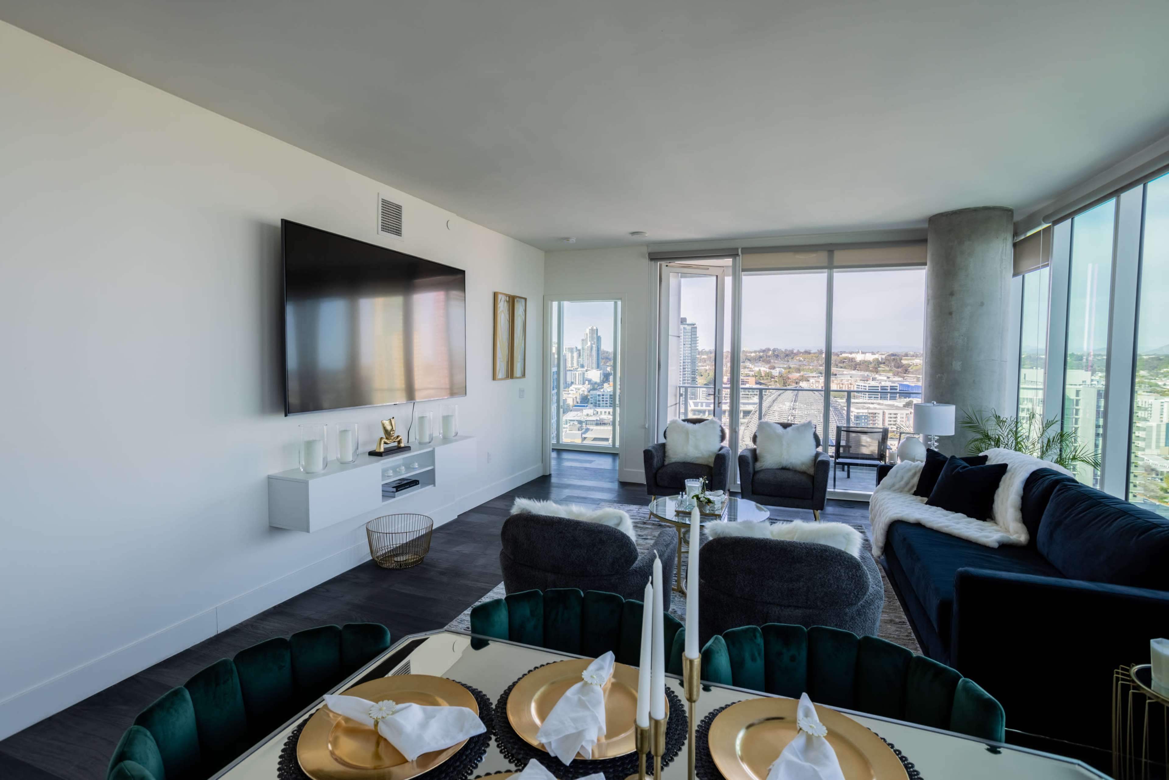 A modern living area with a television mounted on the wall, a dining table set for a meal, and large windows offering a view of the city.