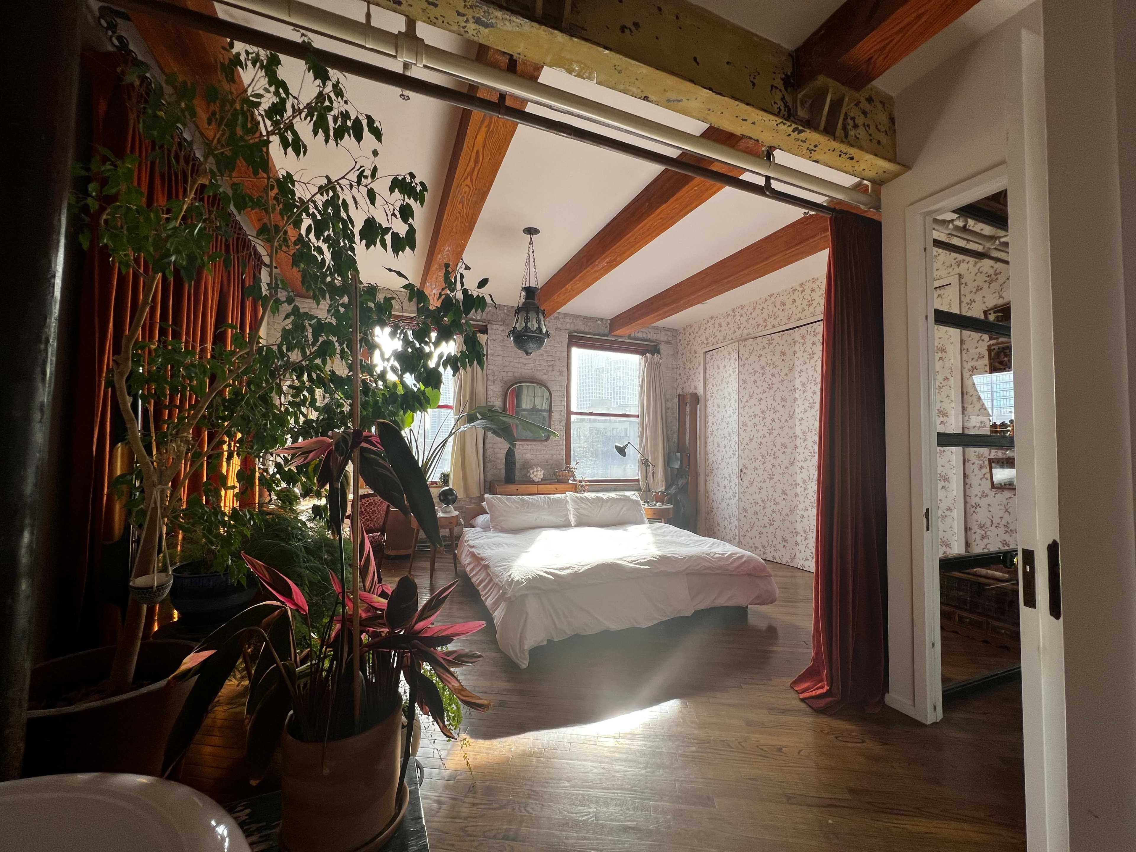 The image shows a cozy bedroom with a large bed, wooden ceiling beams, plants, and soft natural light streaming through the windows.