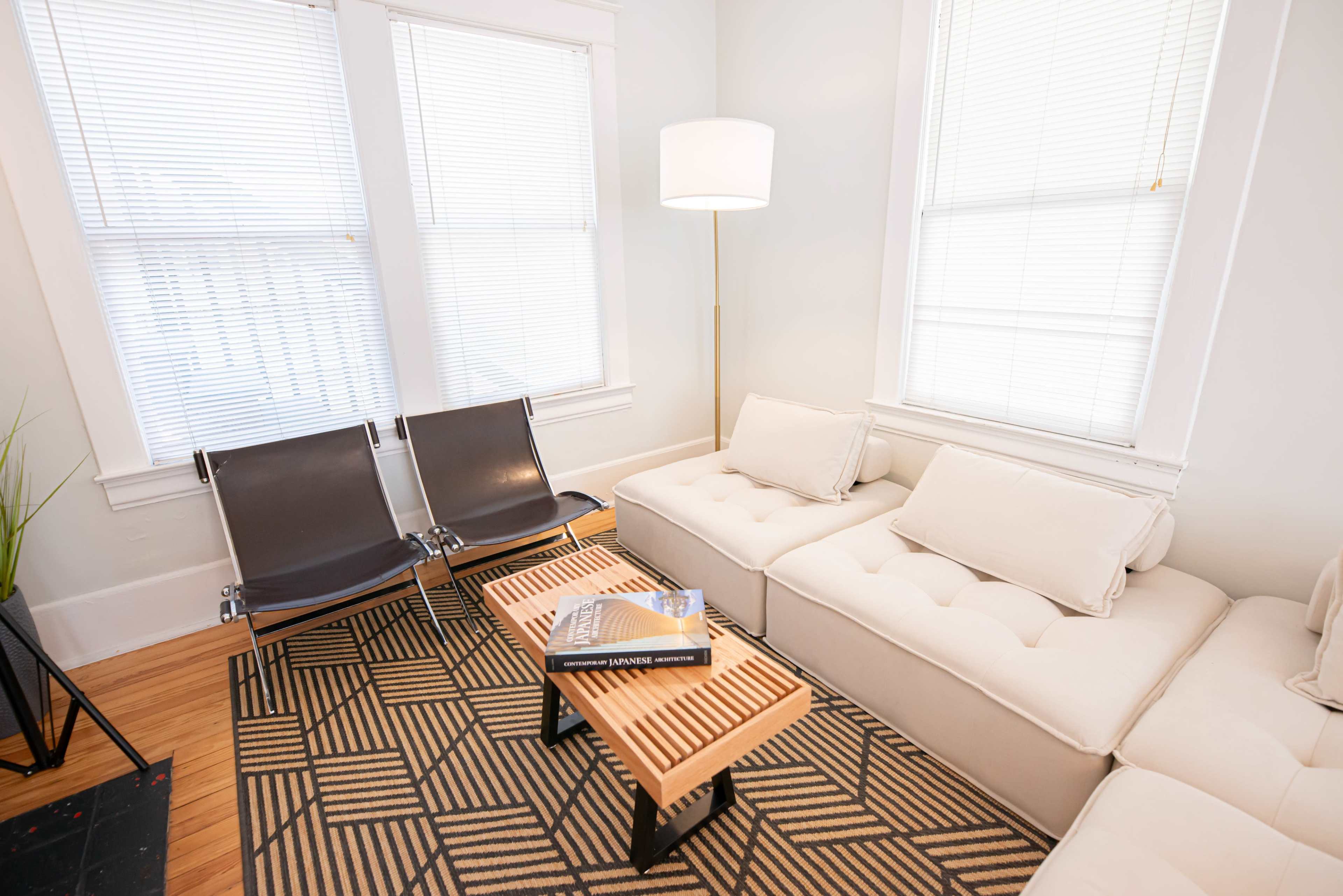 A bright living room with large windows, a sectional sofa, two black chairs, and a wooden coffee table stacked with books.