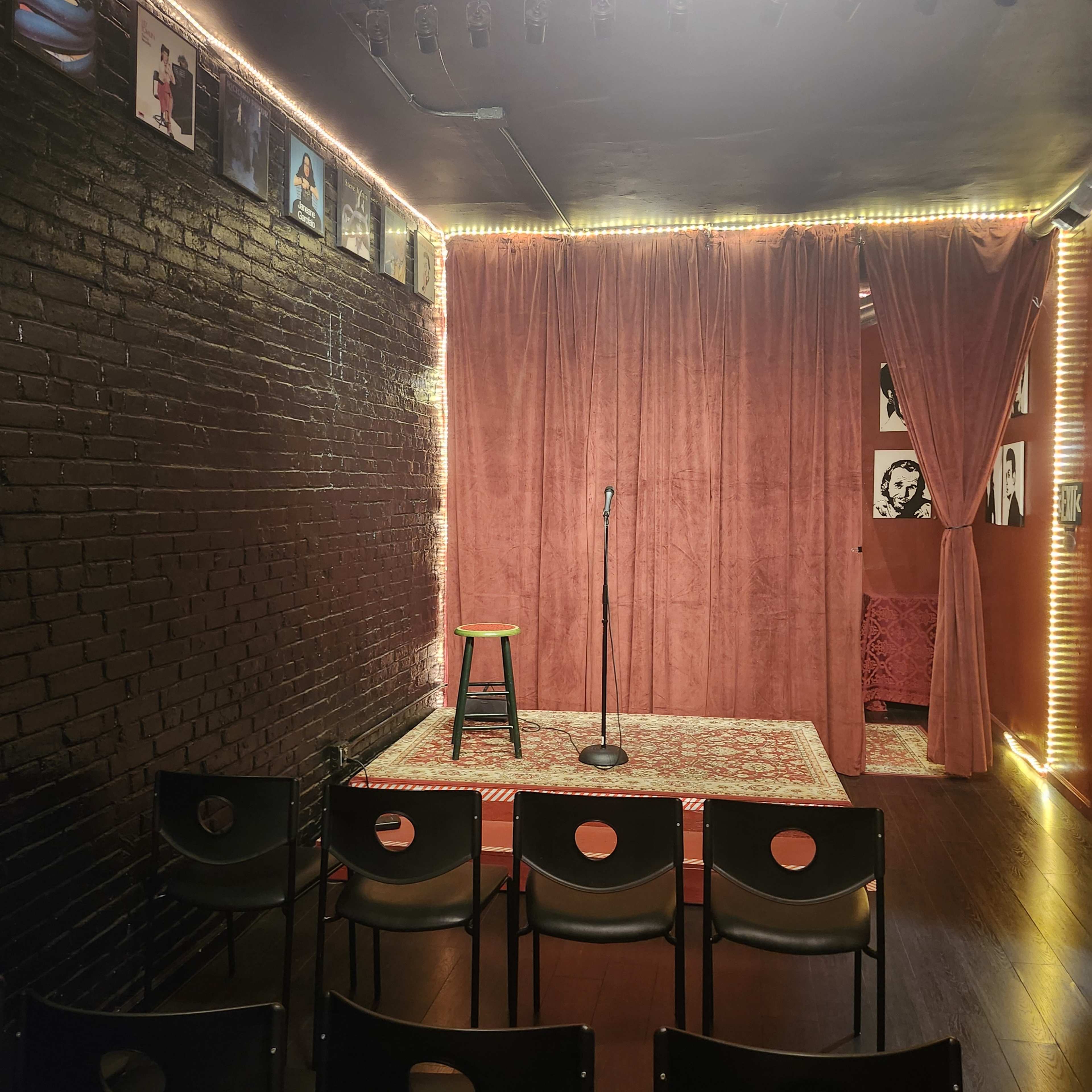 The image shows a small performance space with a microphone stand and a stool on a stage surrounded by red curtains, black brick walls, and illuminated by string lights, with empty chairs arranged in front.