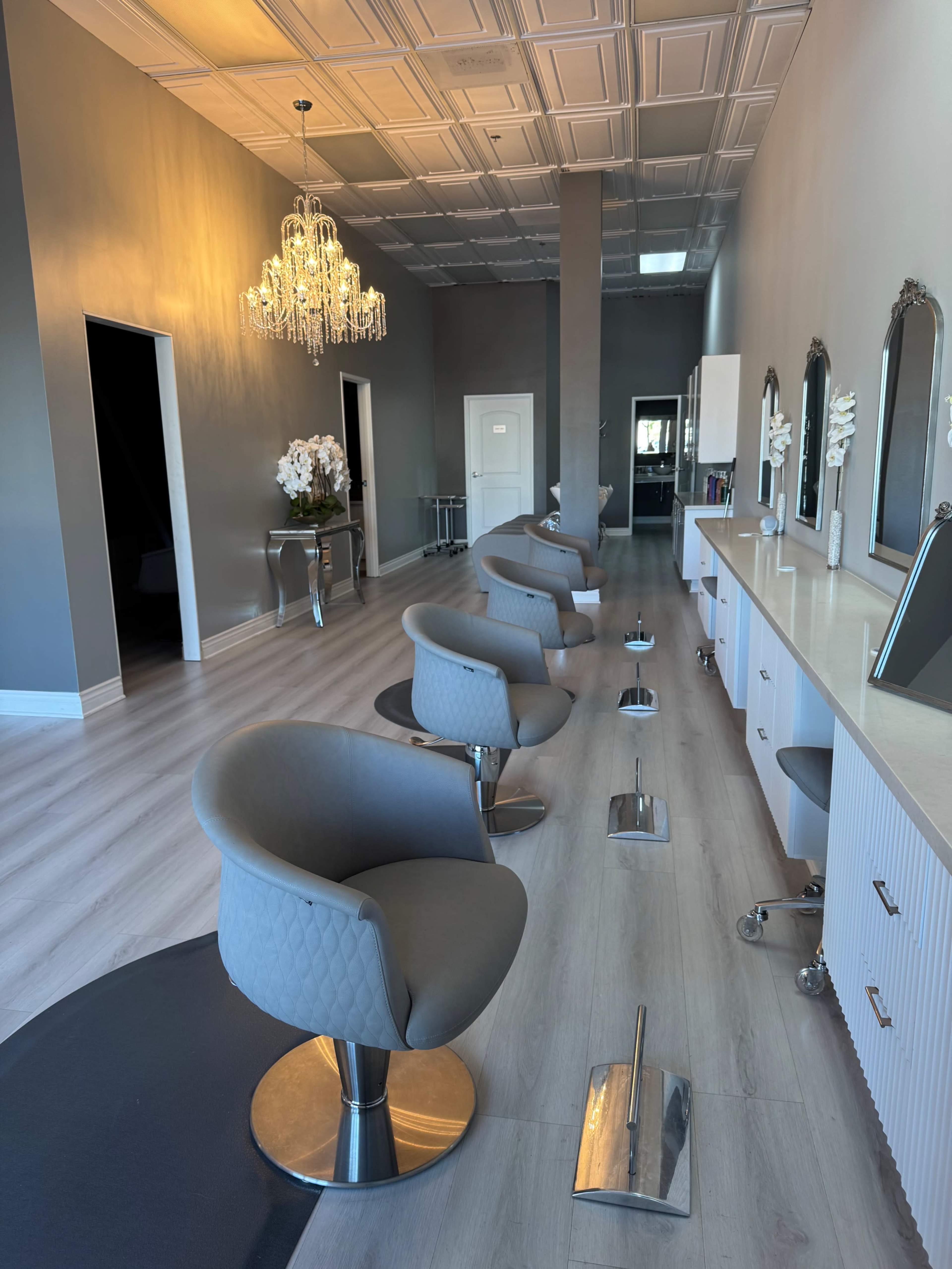 The image shows a modern hair salon interior featuring gray styling chairs arranged along a well-lit, spacious area with mirrors and decorative elements.
