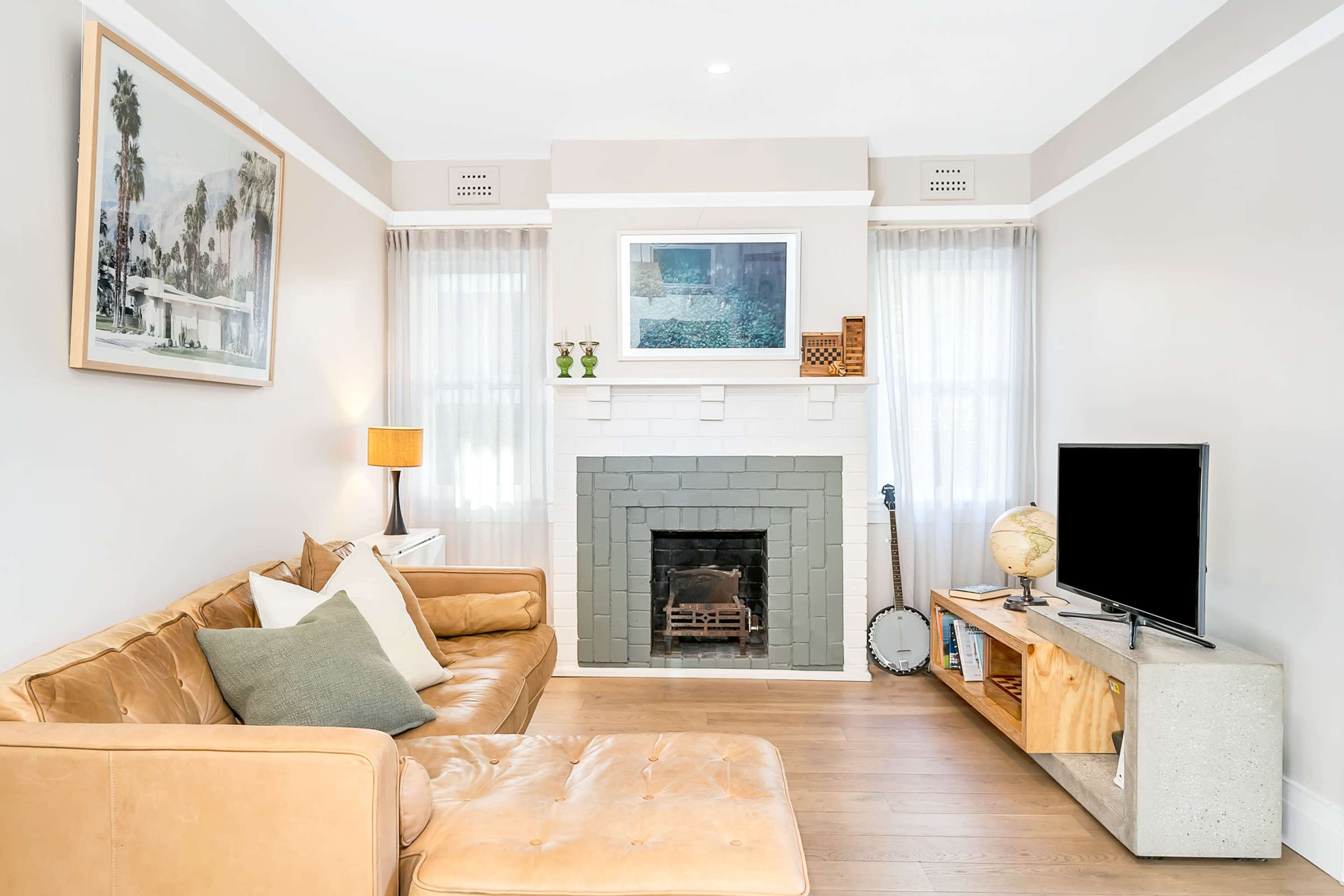 A modern living room featuring a tan leather sofa, a fireplace with a grey brick facade, large windows with sheer curtains, a television on a wooden stand, and a decorative globe near the fireplace.