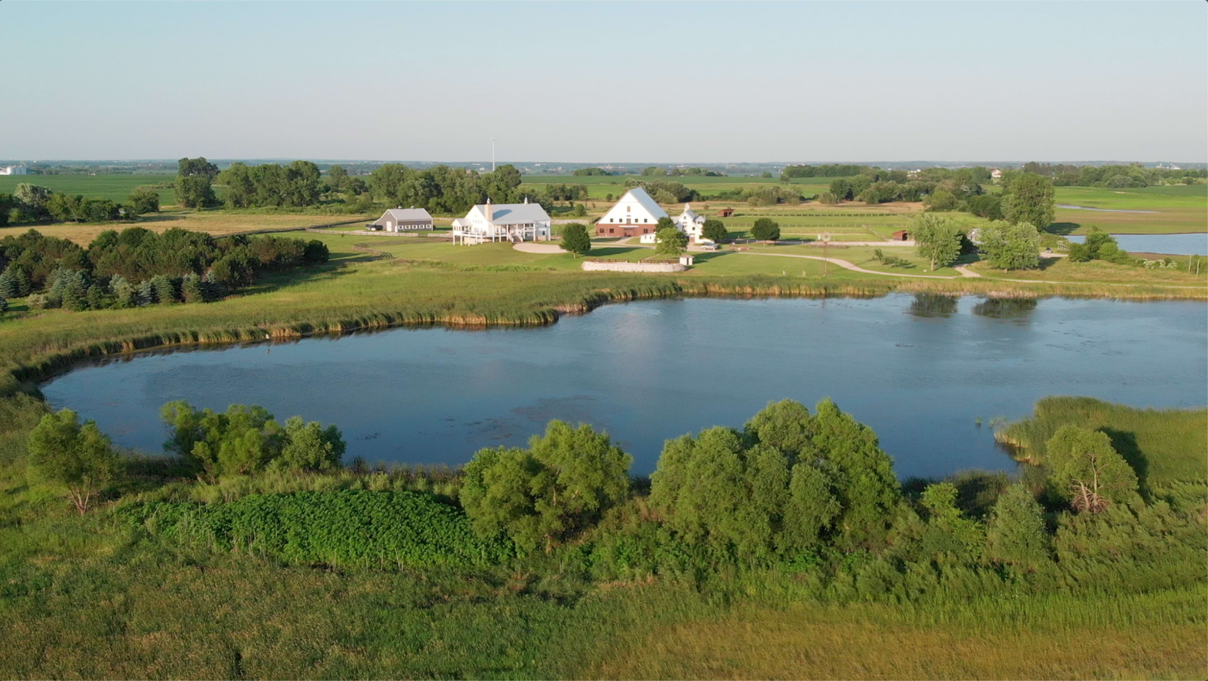 The image shows a peaceful landscape featuring a pond surrounded by green vegetation and a white farmstead in the background.