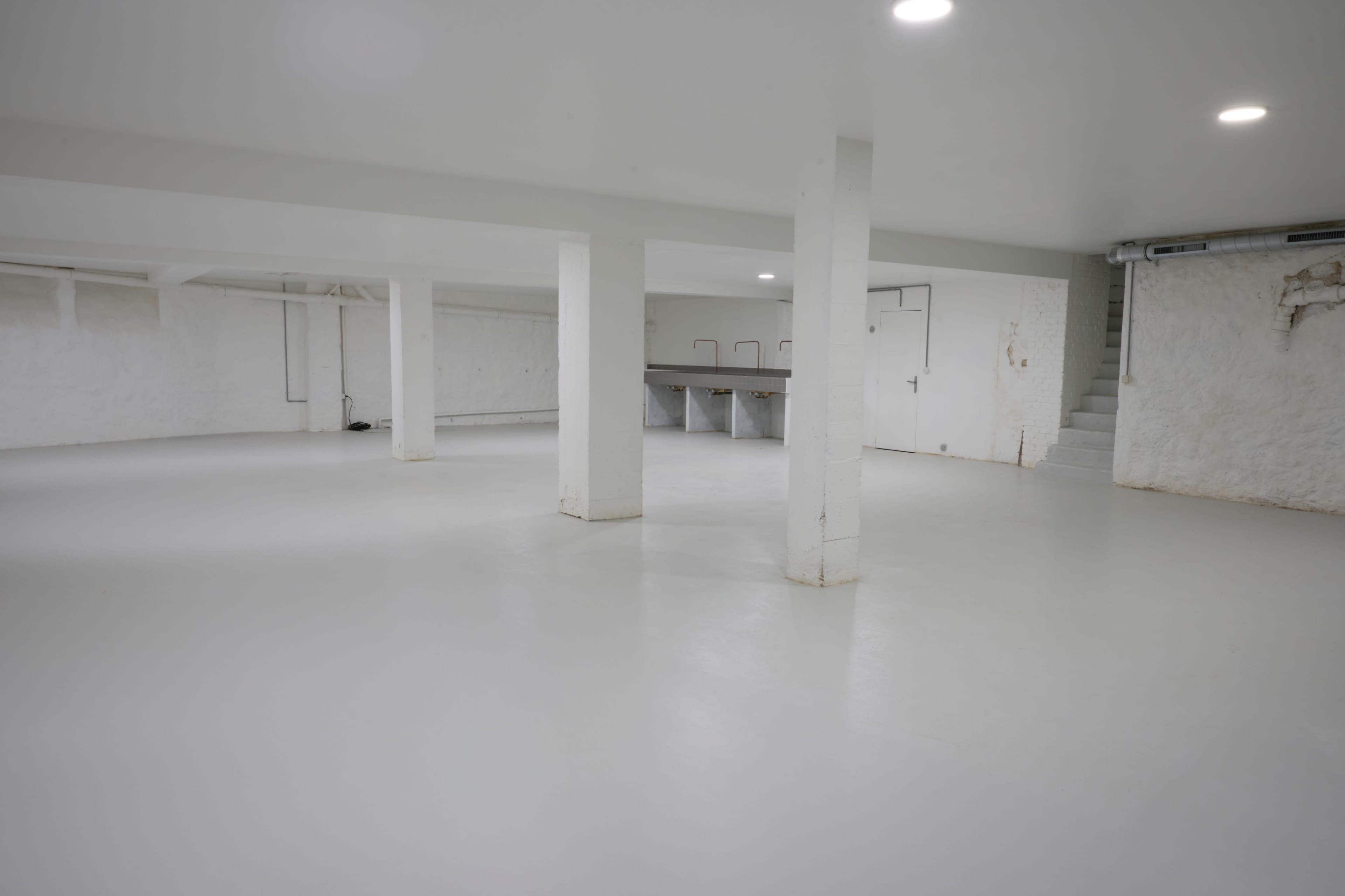 A spacious, empty basement with white walls and a smooth white floor, featuring several support columns and a staircase in one corner.