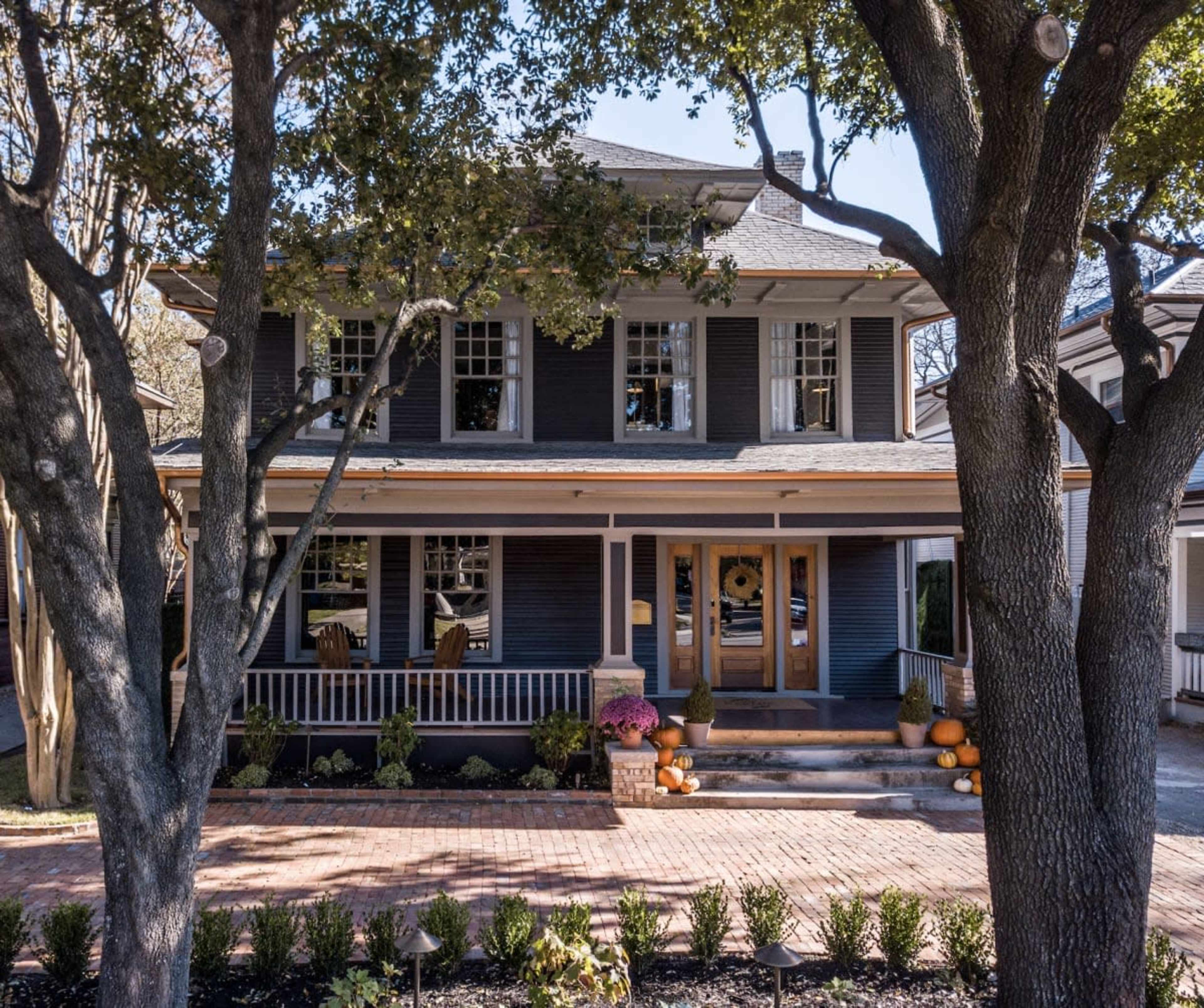 A two-story house with dark blue siding, a covered porch, and pumpkin decorations is surrounded by large trees and manicured landscaping.