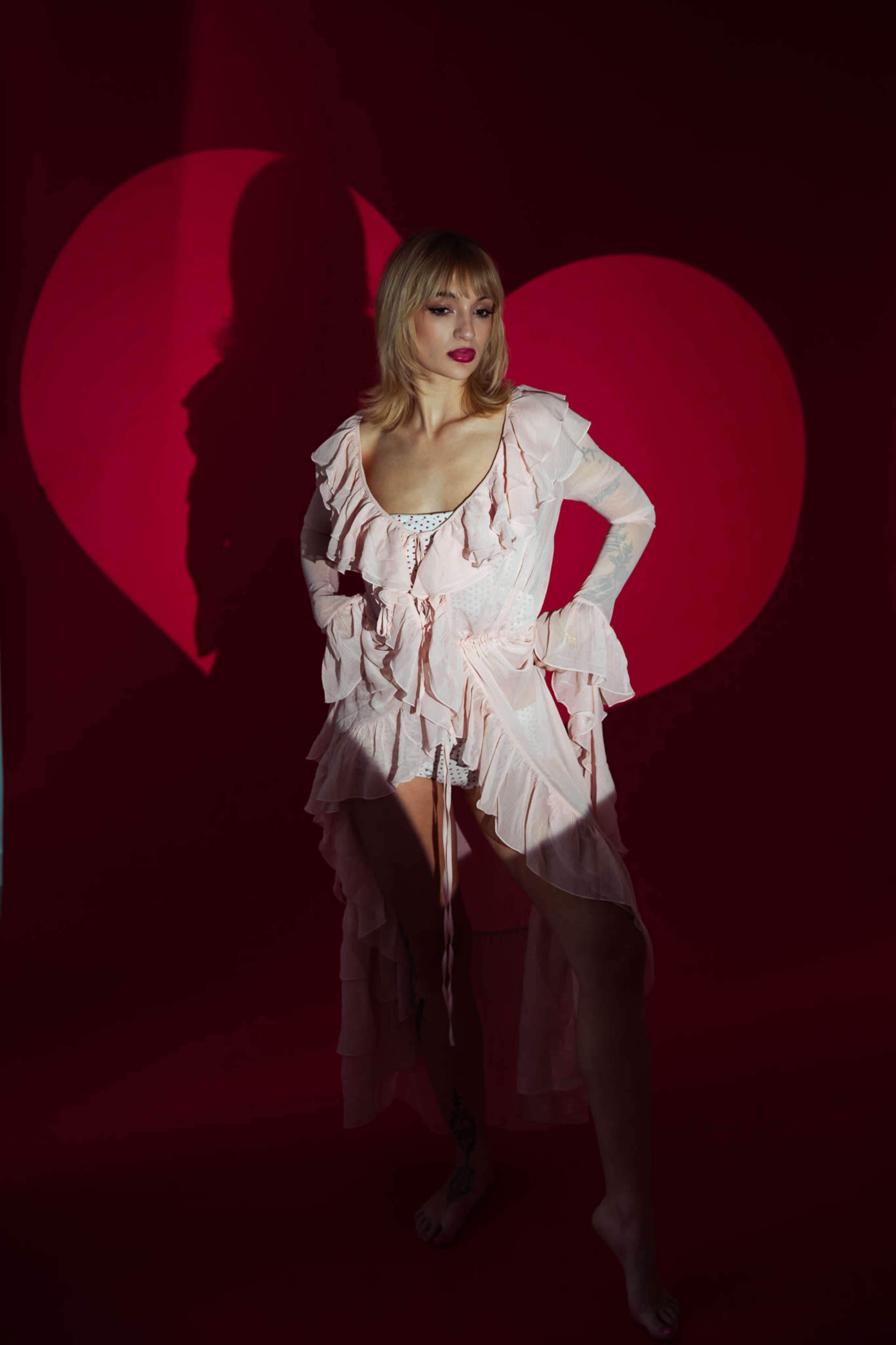 A woman in a pink ruffled dress stands against a red backdrop that features a large heart shadow.
