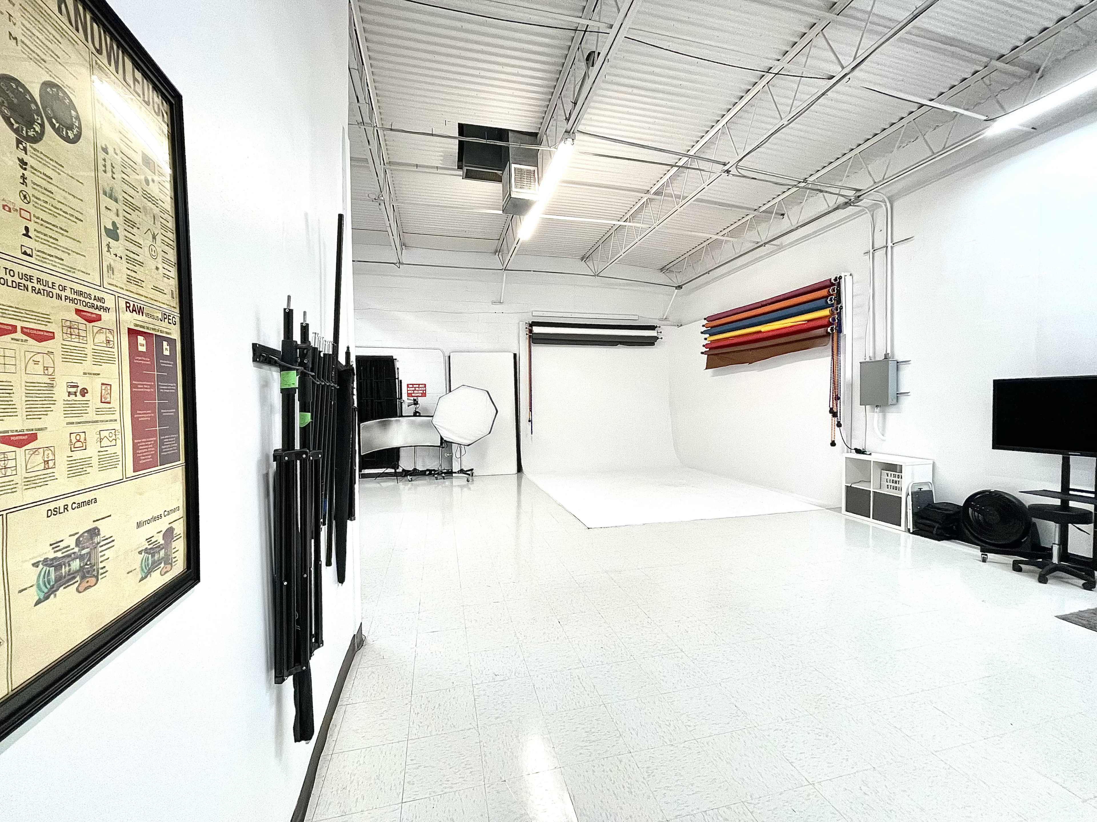 A photography studio with a white backdrop, various colored backdrop rolls neatly hung, and equipment stored along the walls.