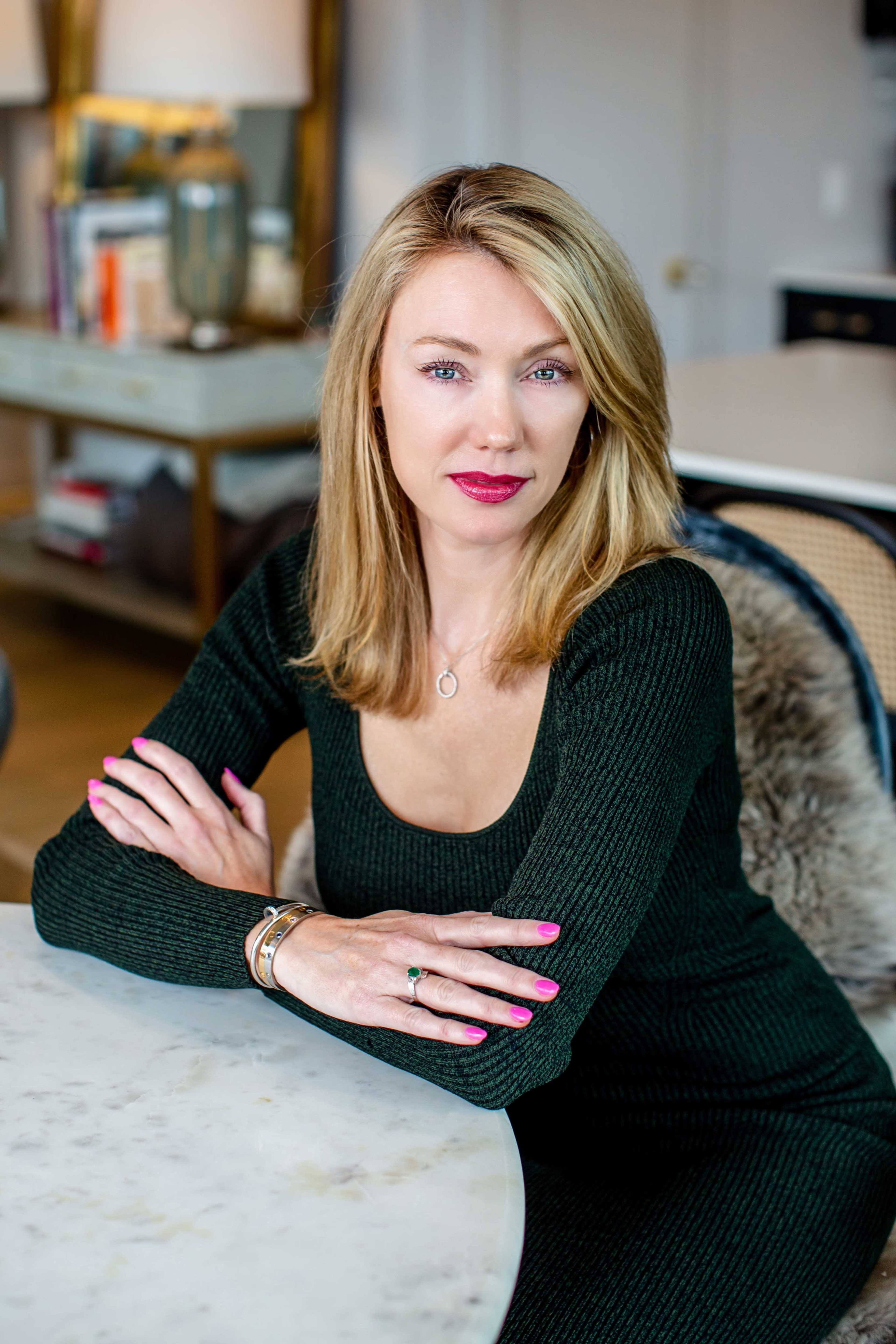 A woman with blonde hair sits at a table, wearing a dark green dress and displaying manicured nails, while resting her arms on the table's surface.