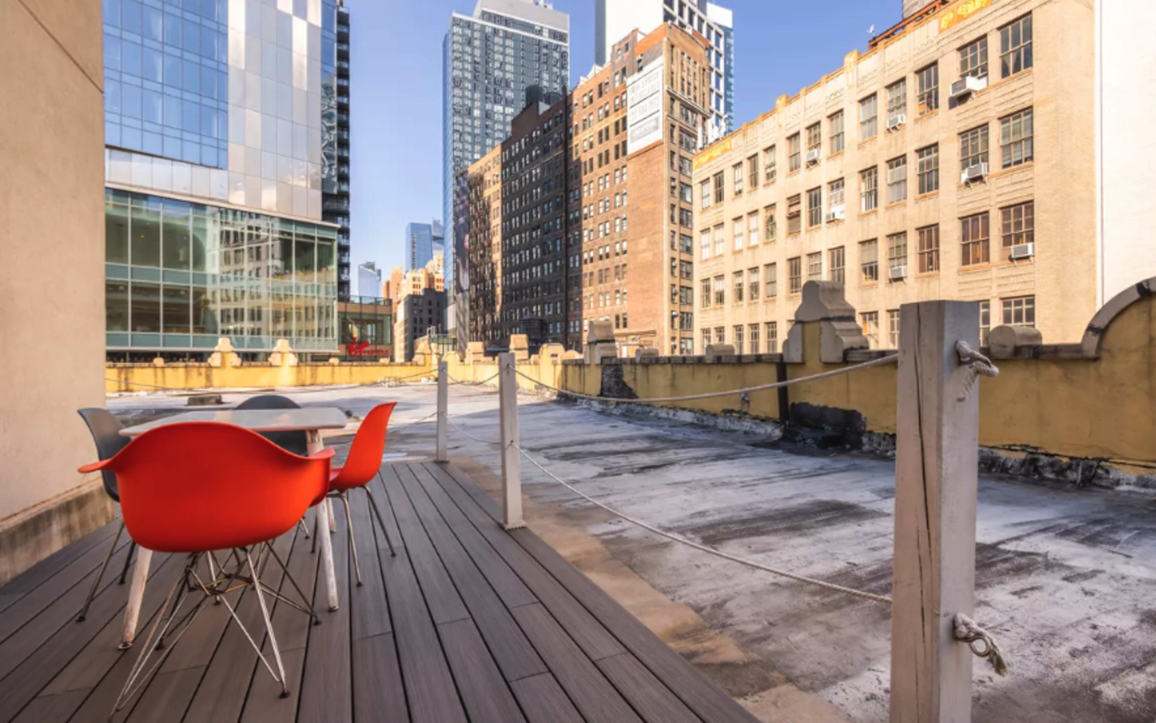 A rooftop terrace features a wooden deck with a table and four chairs, overlooking a cityscape of tall buildings.