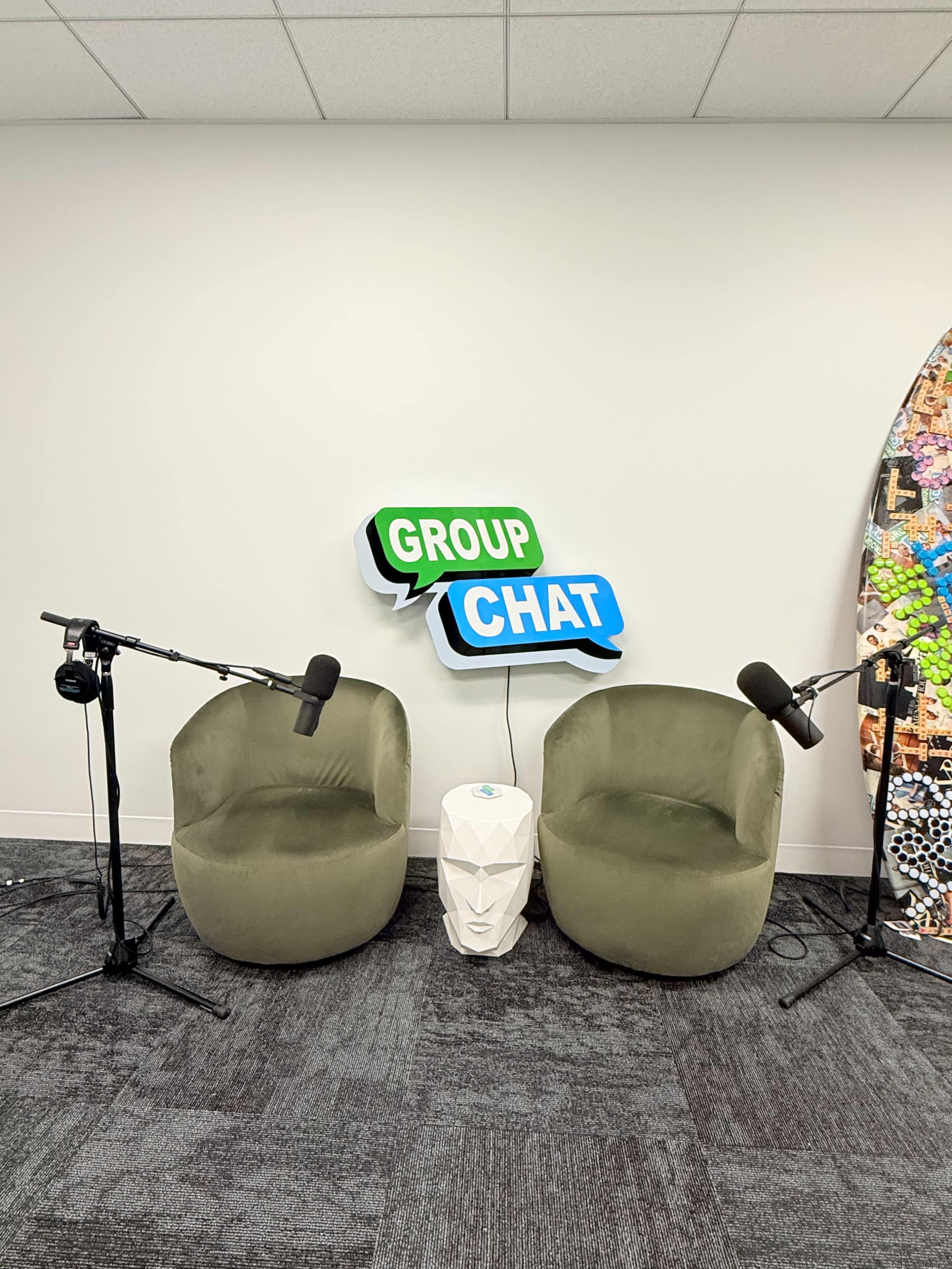 The image shows two green chairs placed facing each other with microphones on stands and a decorative sign that says "GROUP CHAT" in the background.