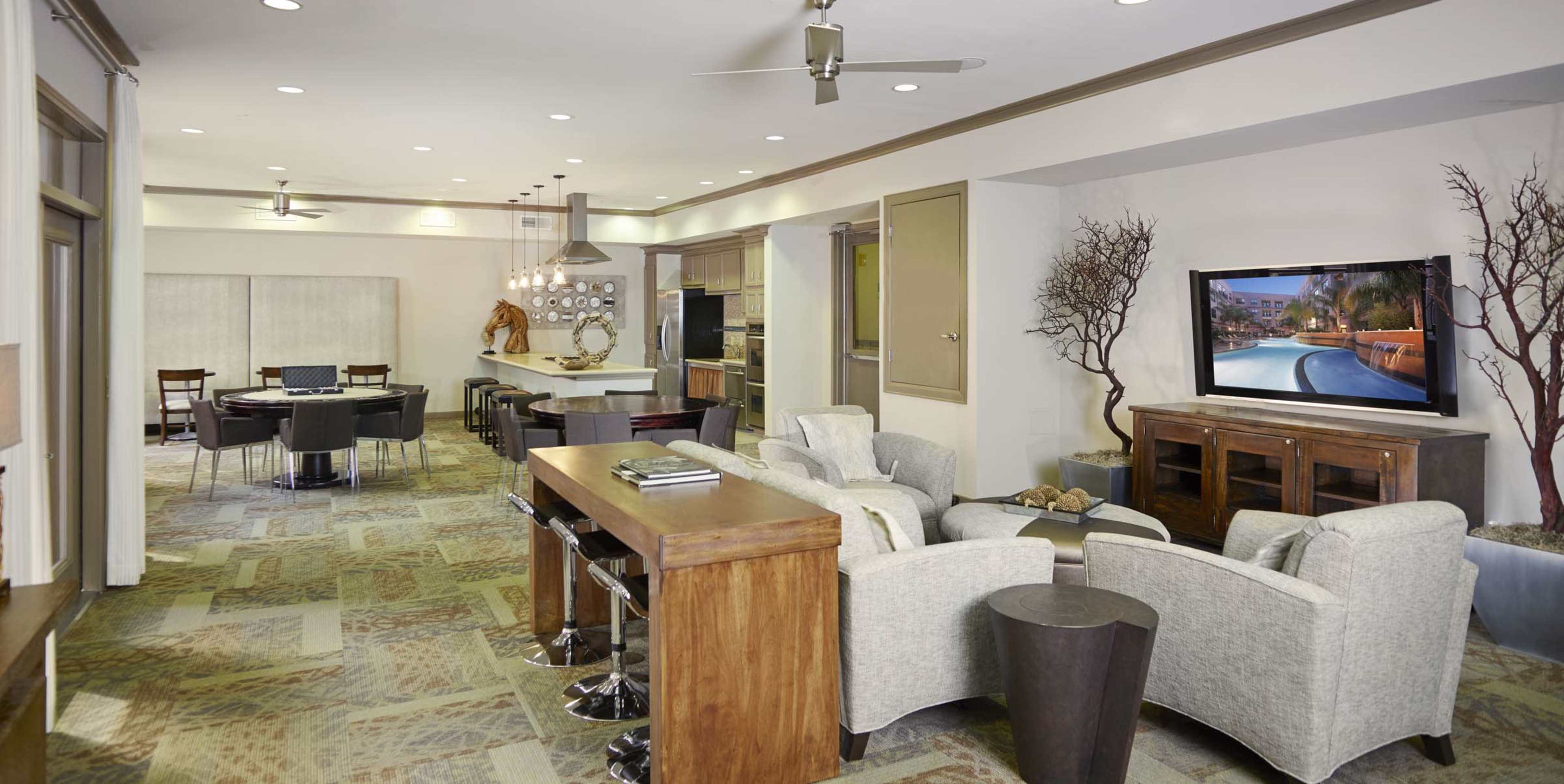 A modern living space with a seating area featuring two light-colored chairs, a wooden bar, and a television mounted on the wall, adjacent to a dining area with a round table and several chairs.
