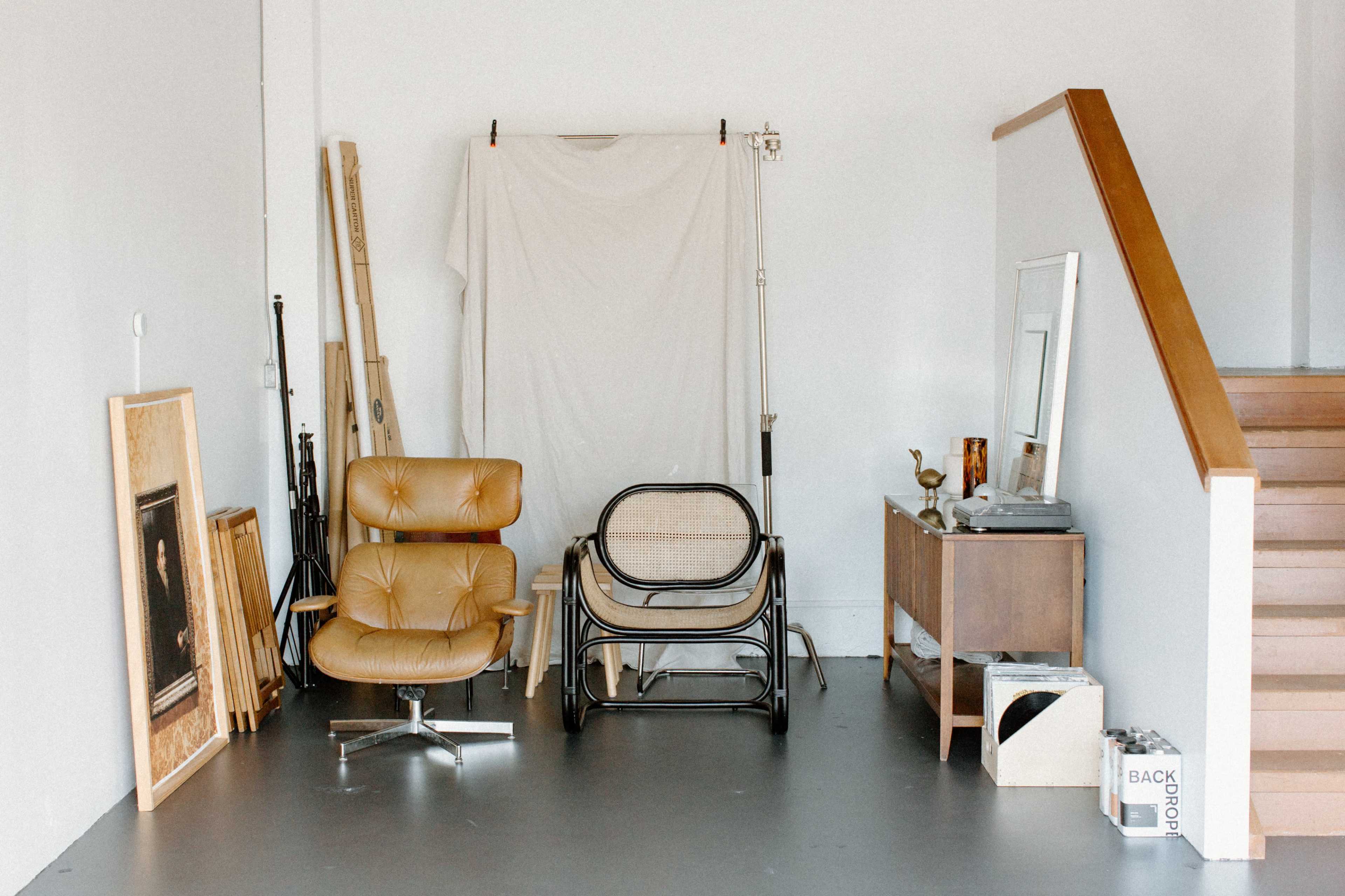 A minimalist interior with two chairs, a wooden staircase, a mirror, and various pieces of artwork and decor arranged against a light-colored wall.