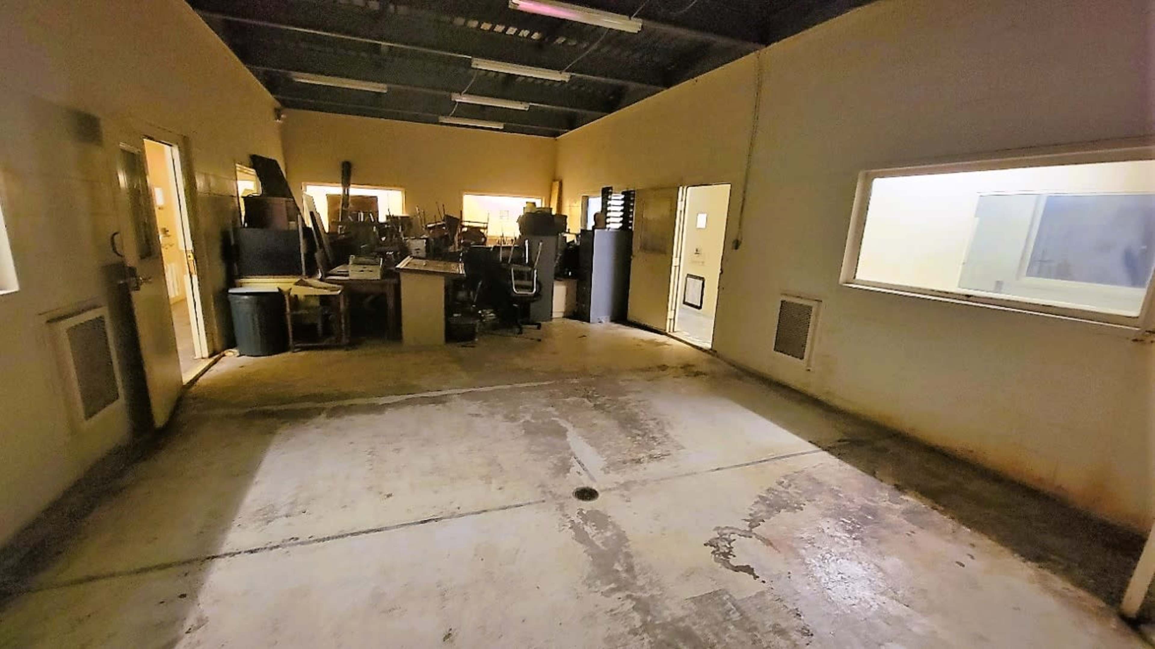 A dimly lit, empty room with concrete flooring and two glass-paneled doors leading to adjacent areas filled with various equipment and furniture.
