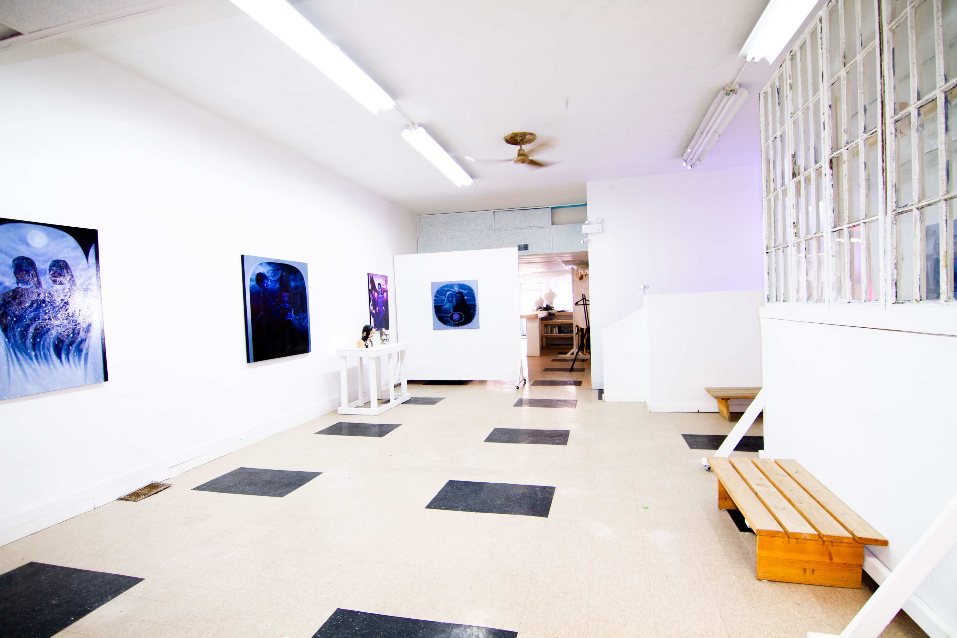 The image shows a minimalist art gallery with white walls, black and white tiled flooring, and two framed artworks displayed on the walls.