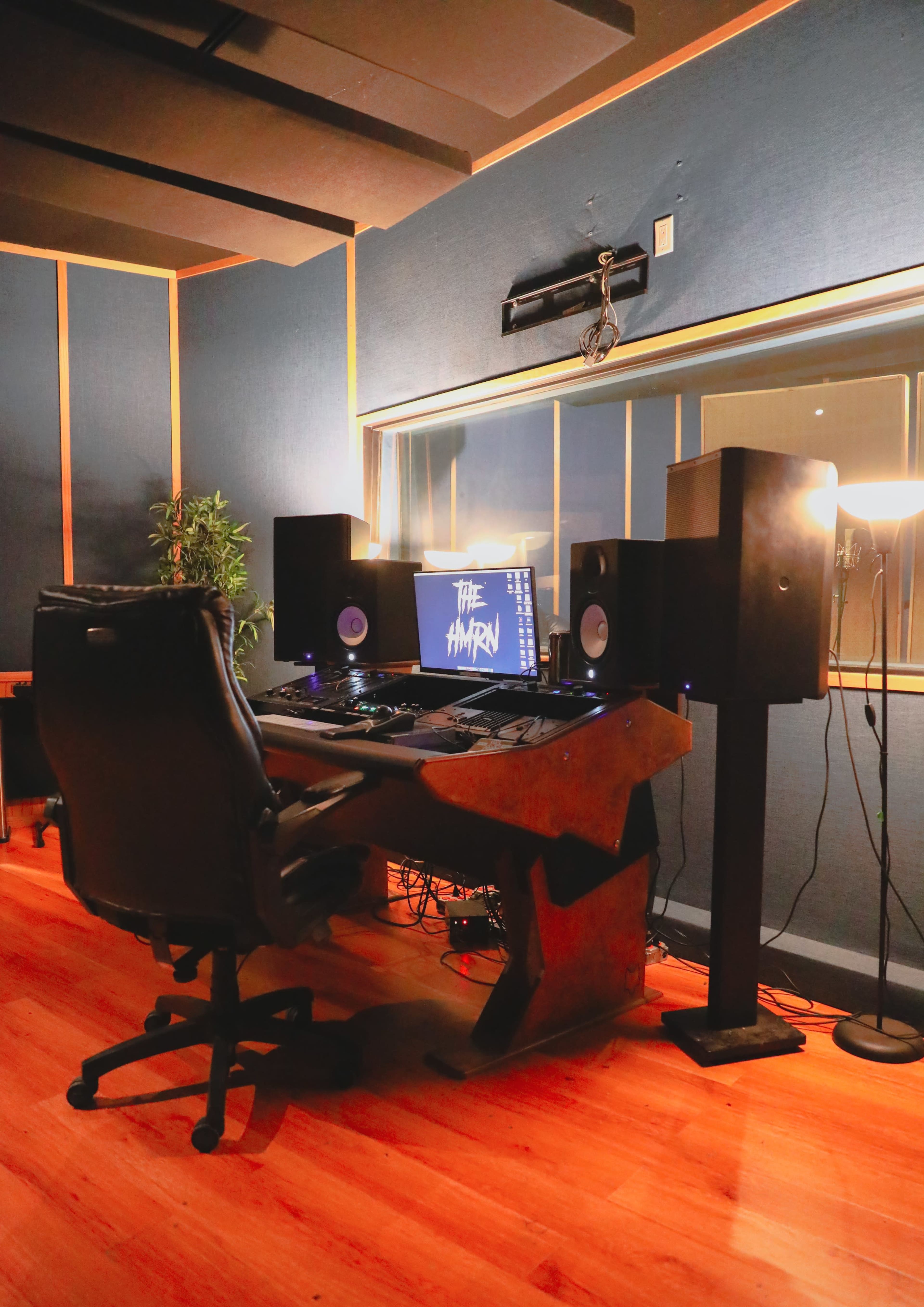The image shows a music studio with a mixing console, speakers, and a computer monitor inside a room featuring wood flooring and blue walls.