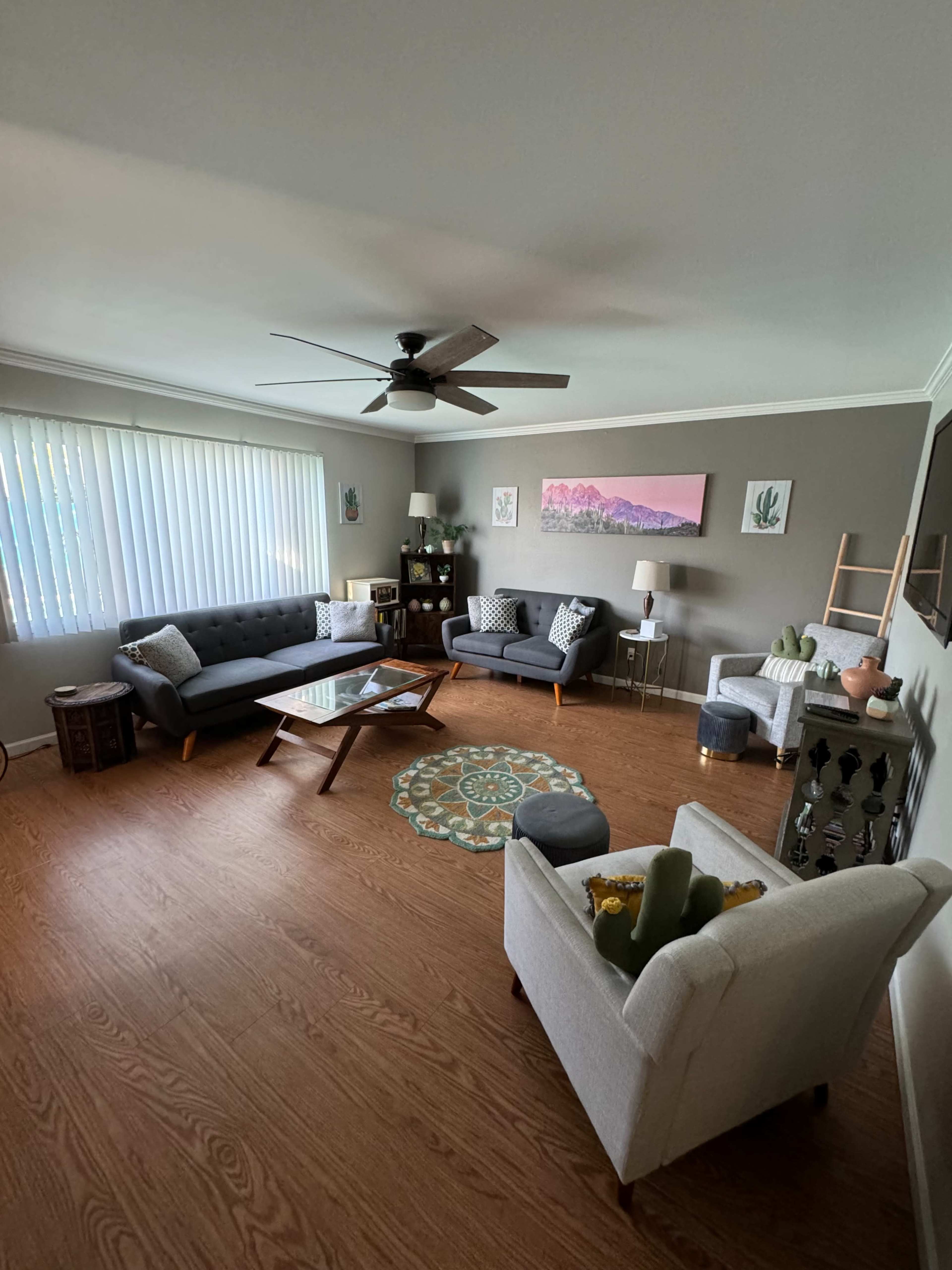 A living room features two gray sofas, a glass coffee table, a ceiling fan, and decorative items, with a large window allowing natural light.