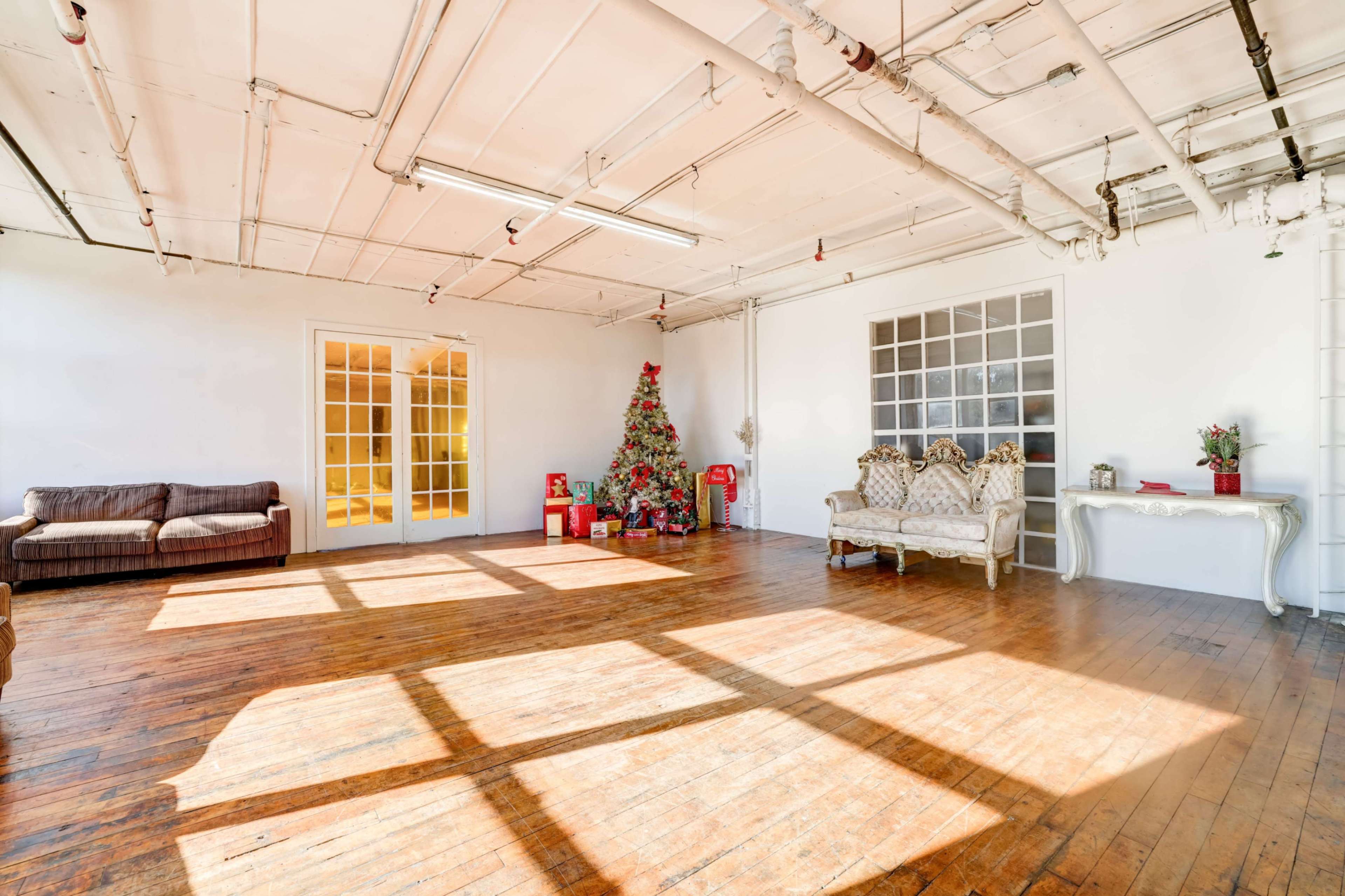 The image shows a spacious, well-lit room with wooden flooring, featuring a Christmas tree and minimal furniture against white walls.