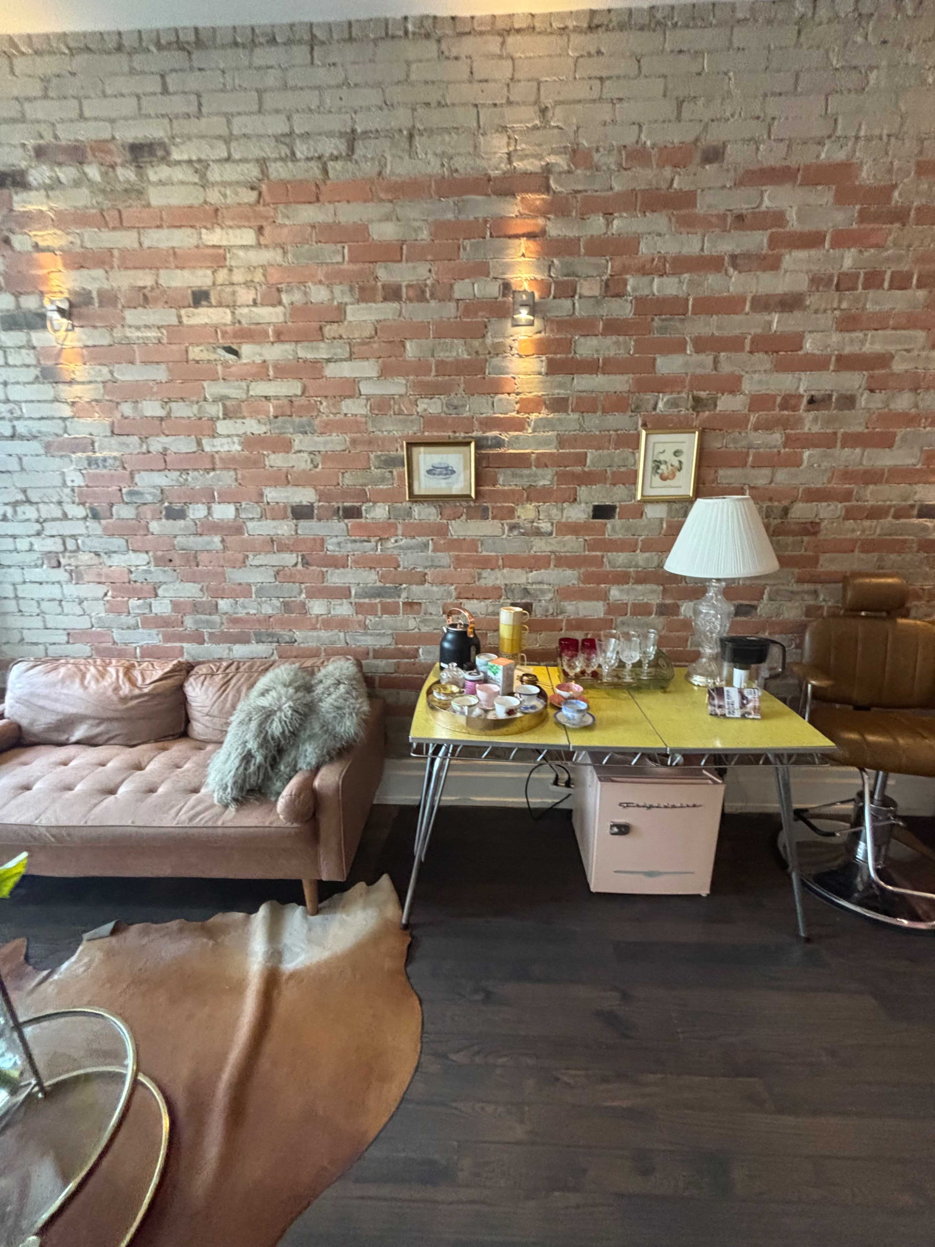 The image shows a cozy interior with a brick wall, a beige sofa, a table set with various items, and a warm light fixture.
