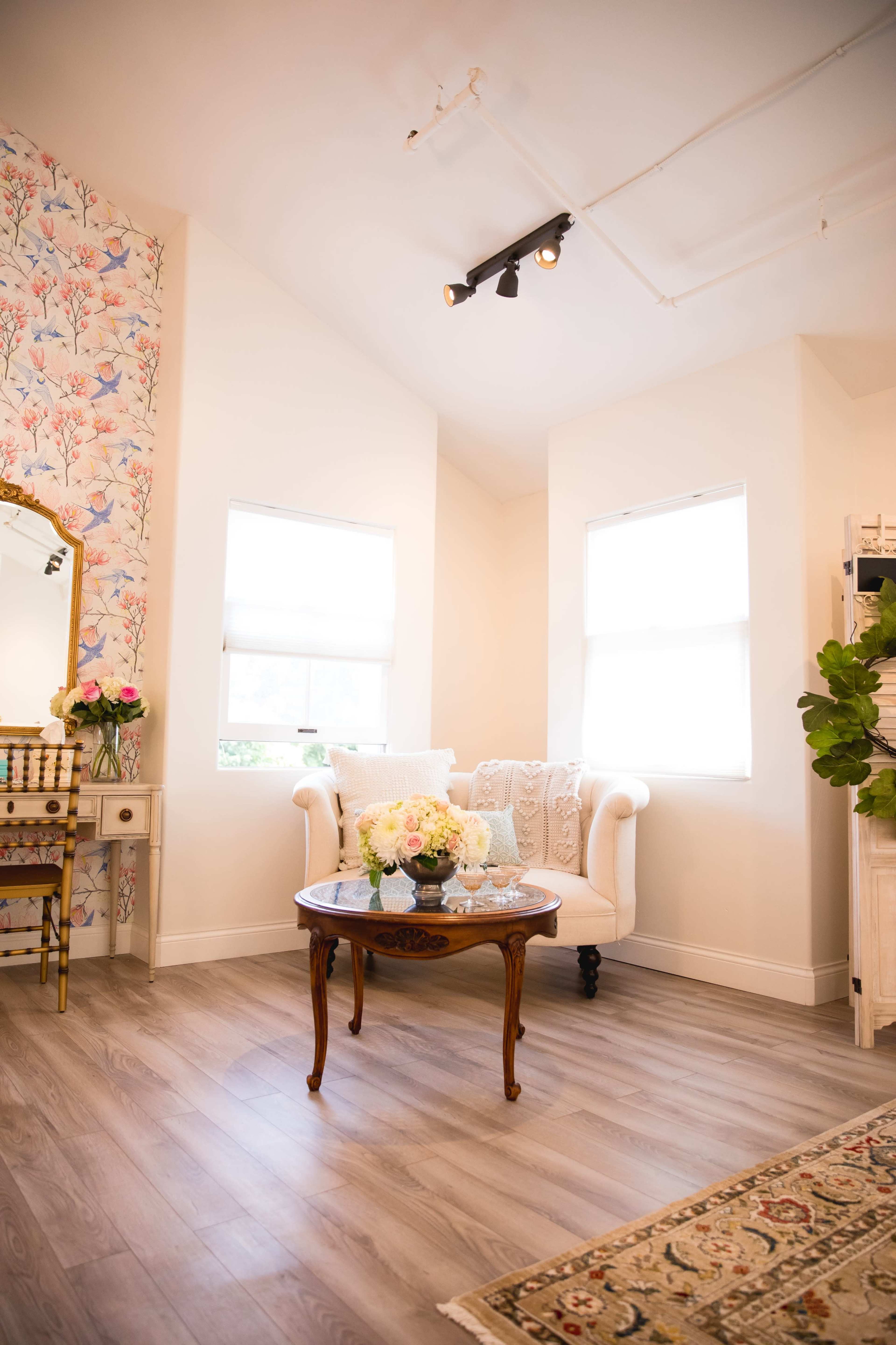 A bright and airy room with a floral wallpapered wall, a vintage-style table with a vase of flowers, and a white upholstered armchair.