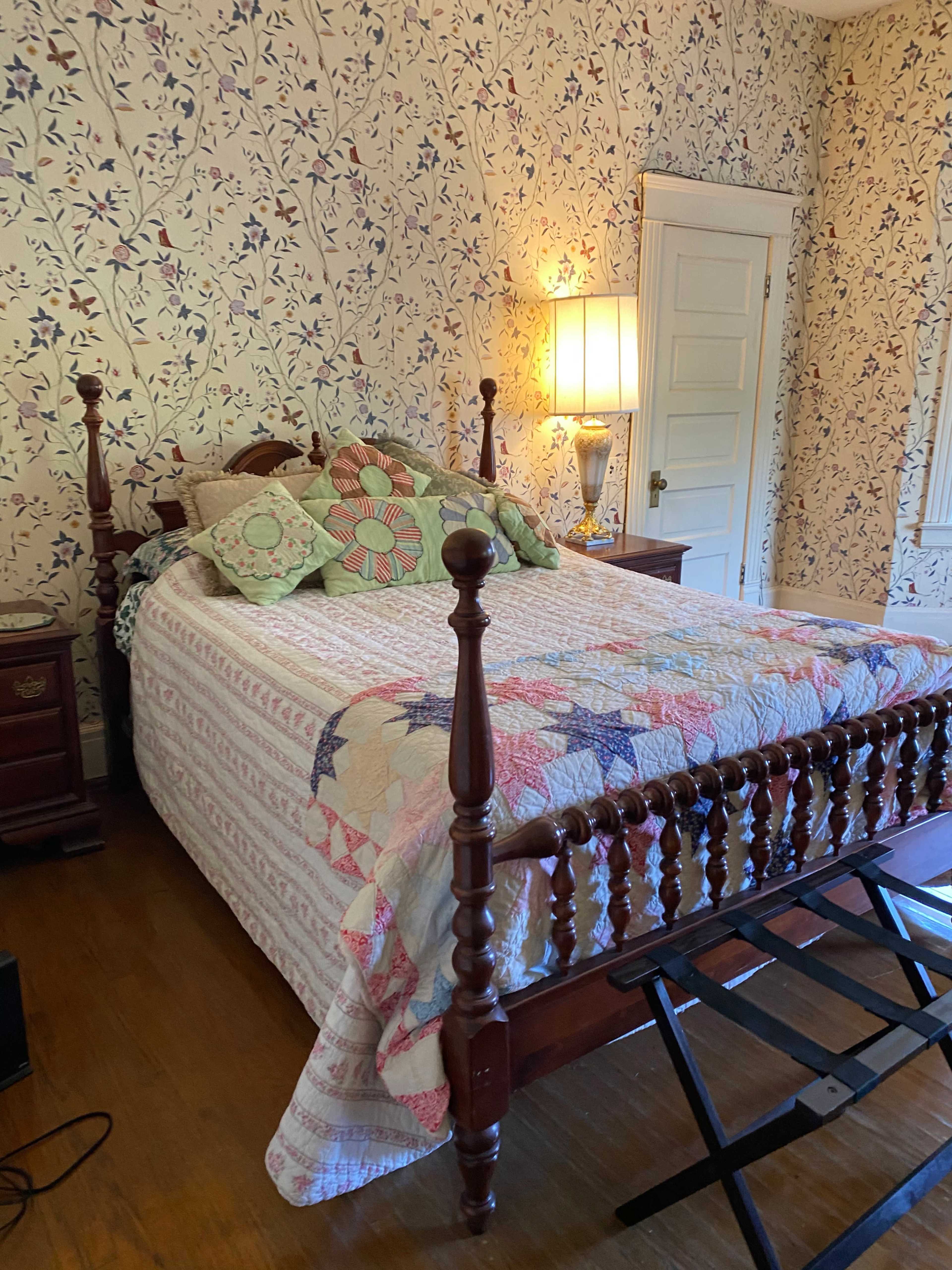 A cozy bedroom with a wooden four-poster bed covered in a colorful quilt, surrounded by floral wallpaper and a bedside table.