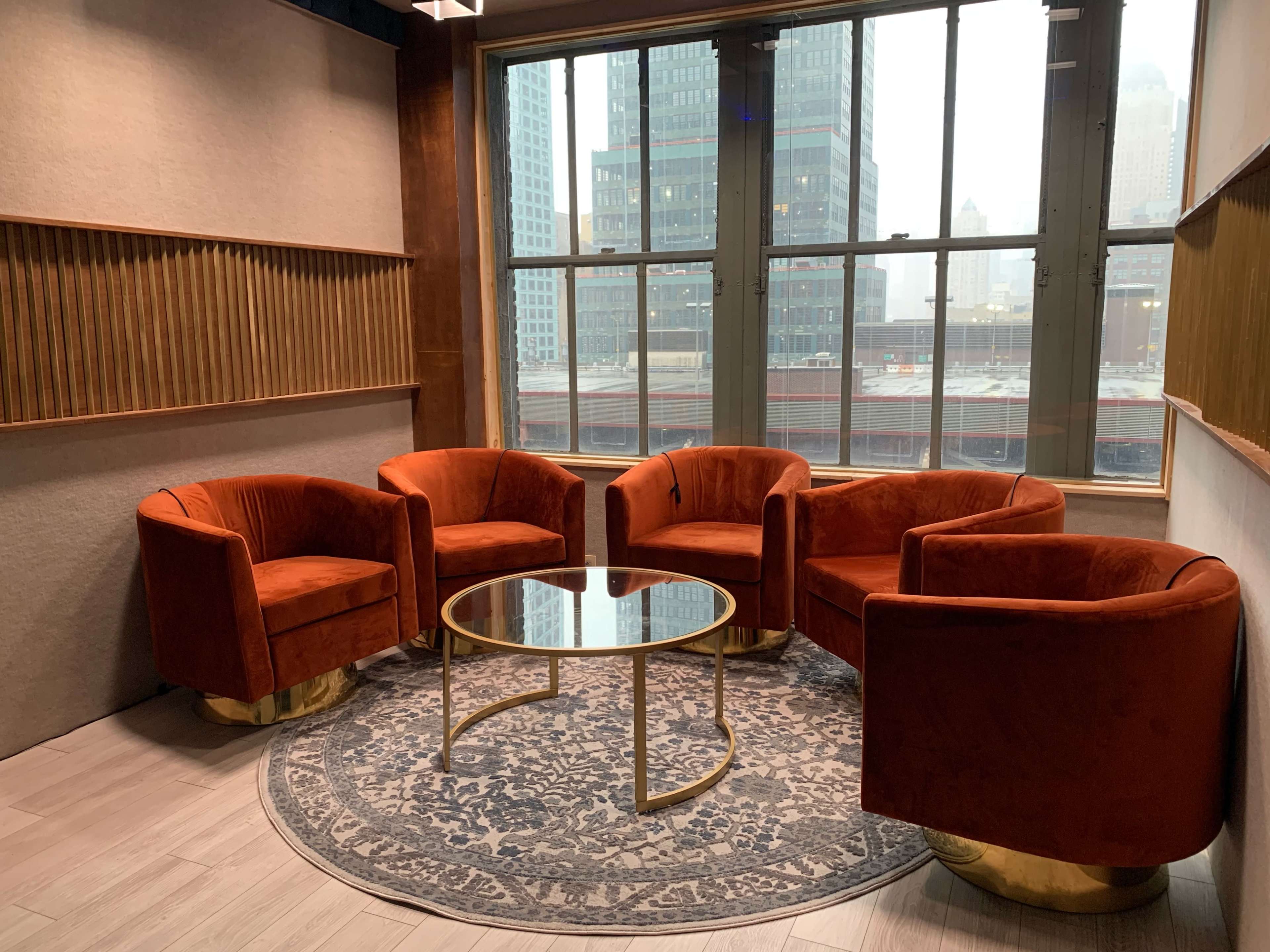 A seating area features four orange velvet chairs arranged around a glass coffee table on a rug in front of large windows overlooking a cityscape.