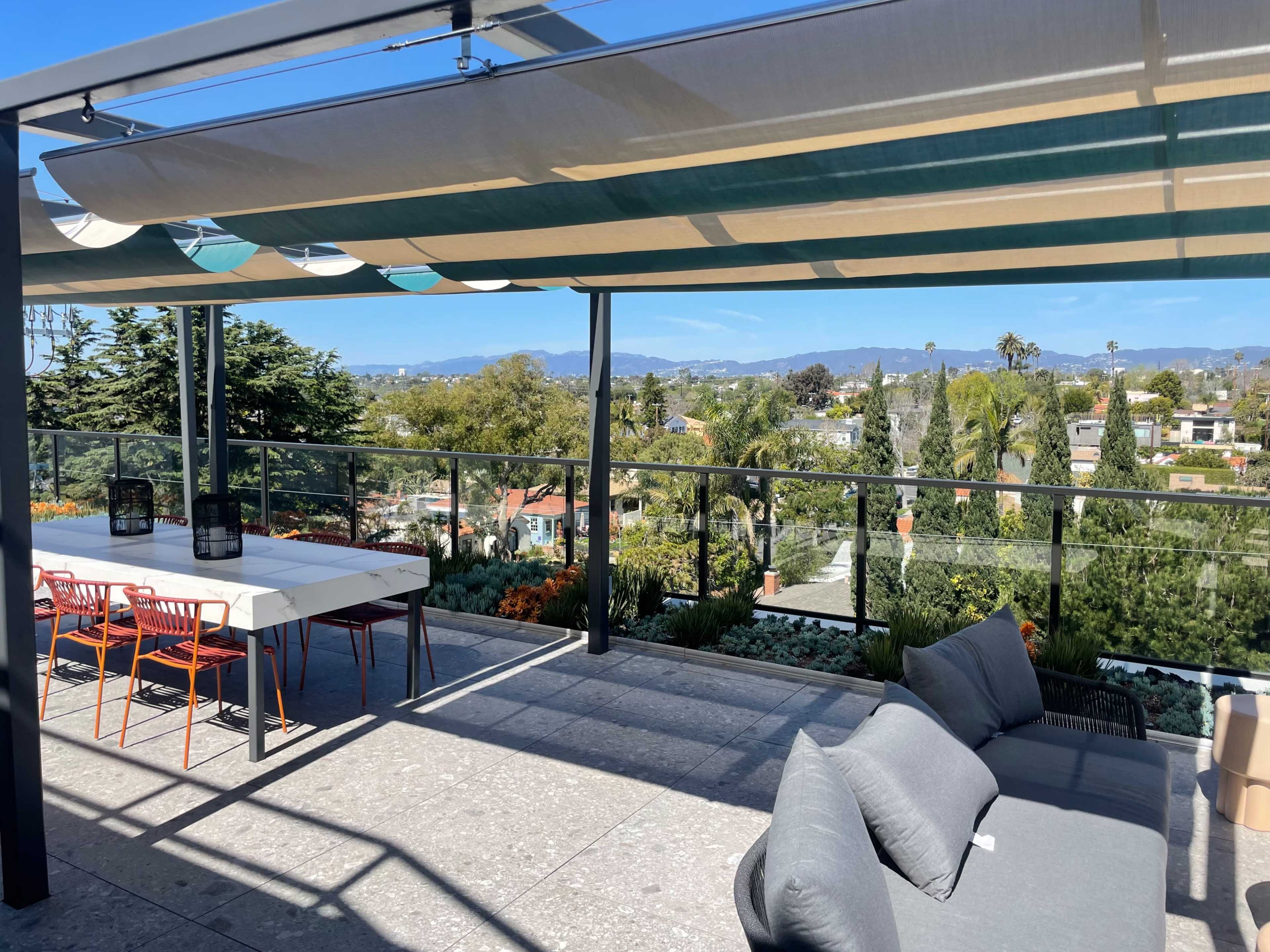 A patio features a table with orange chairs and a gray sofa under a canopy, overlooking a landscape of trees and houses with mountains in the distance.