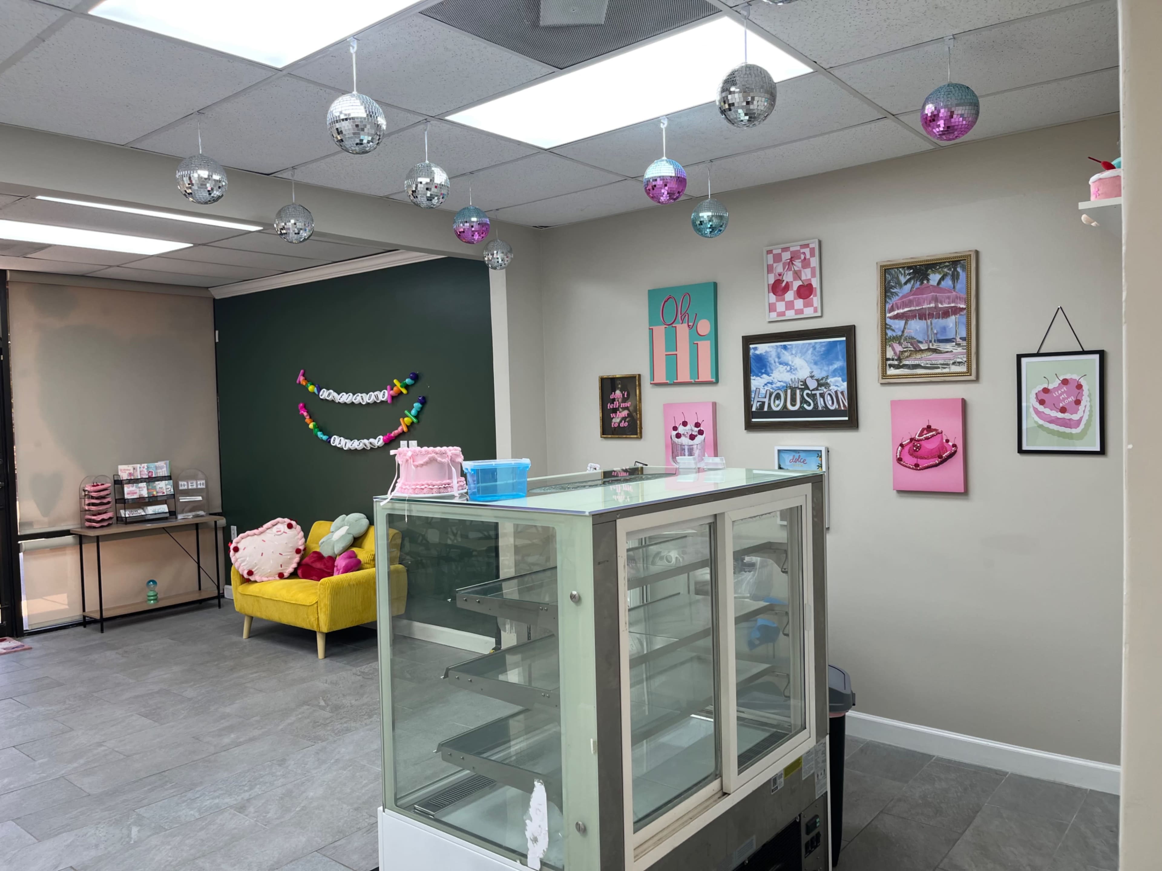 The image shows a brightly decorated dessert shop with a glass display case, colorful wall art, and a yellow couch in the corner.