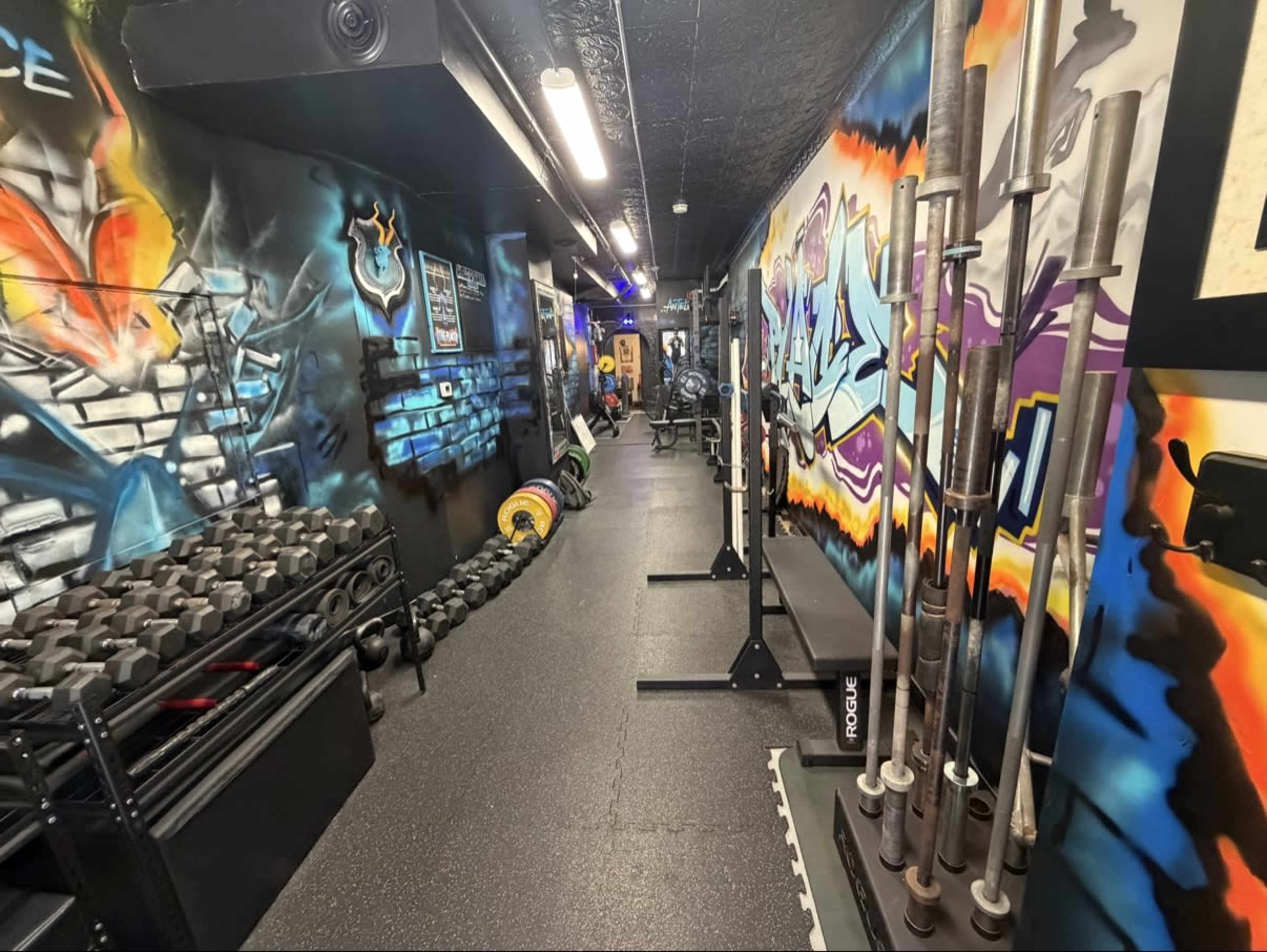 The image shows a narrow gym area featuring colorful graffiti on the walls, with organized rows of weights and various types of exercise equipment along the sides.