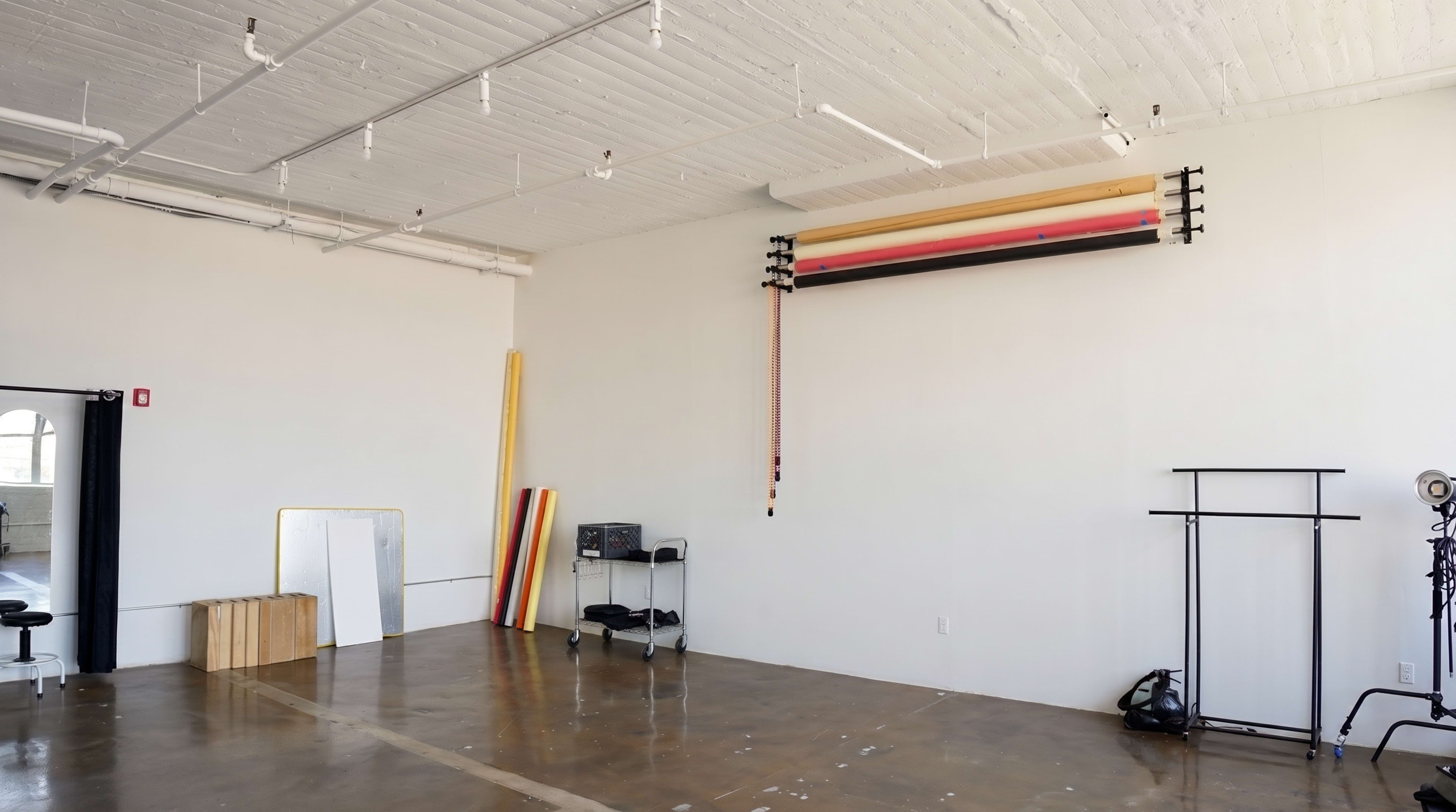 The image shows a spacious, minimalist studio featuring a wall-mounted backdrop system, a large mirror, and various equipment on the floor.