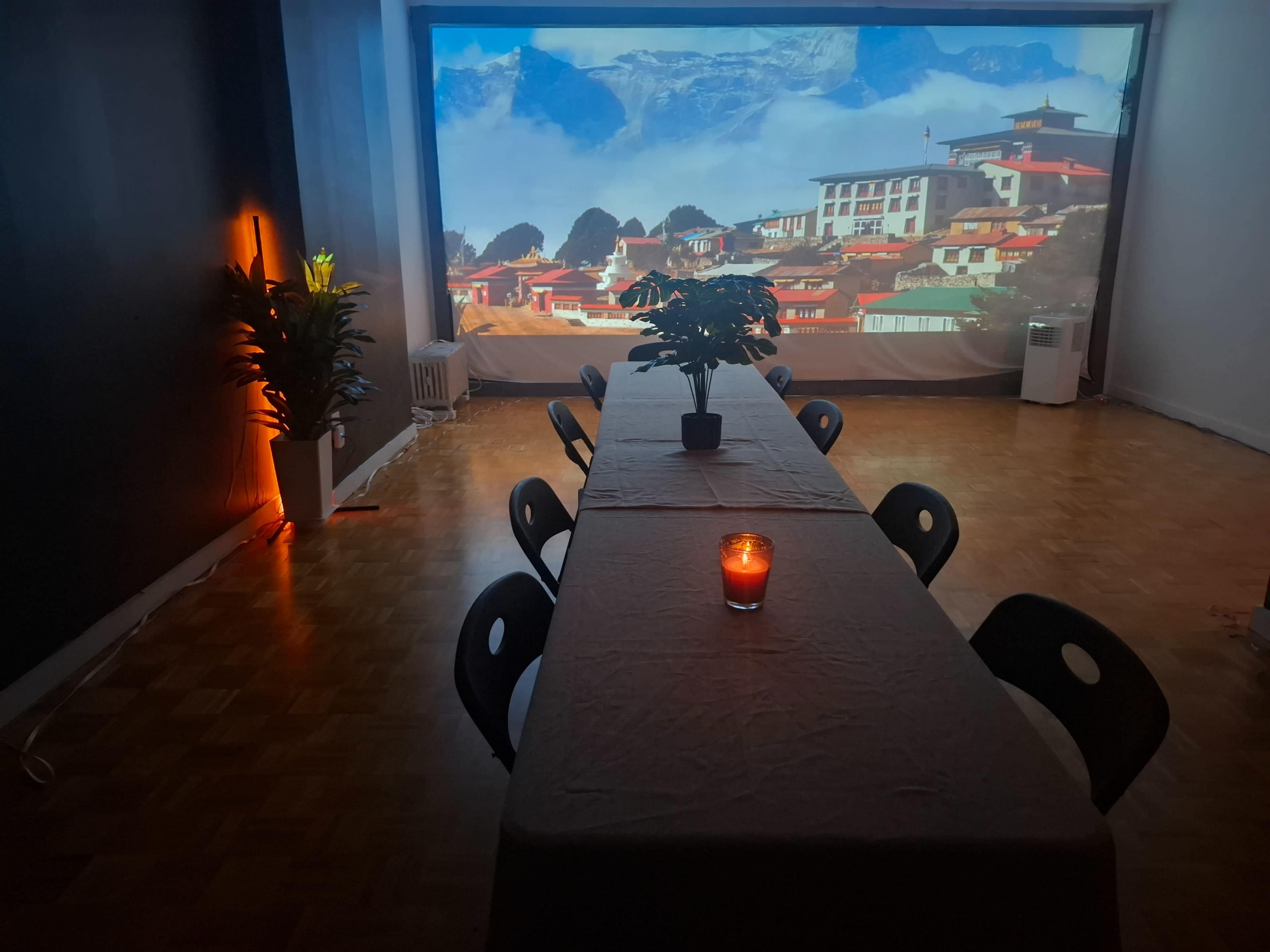 A long table is set up in a spacious room with a large projection of a mountainous landscape on the wall, accompanied by a candle in the center.