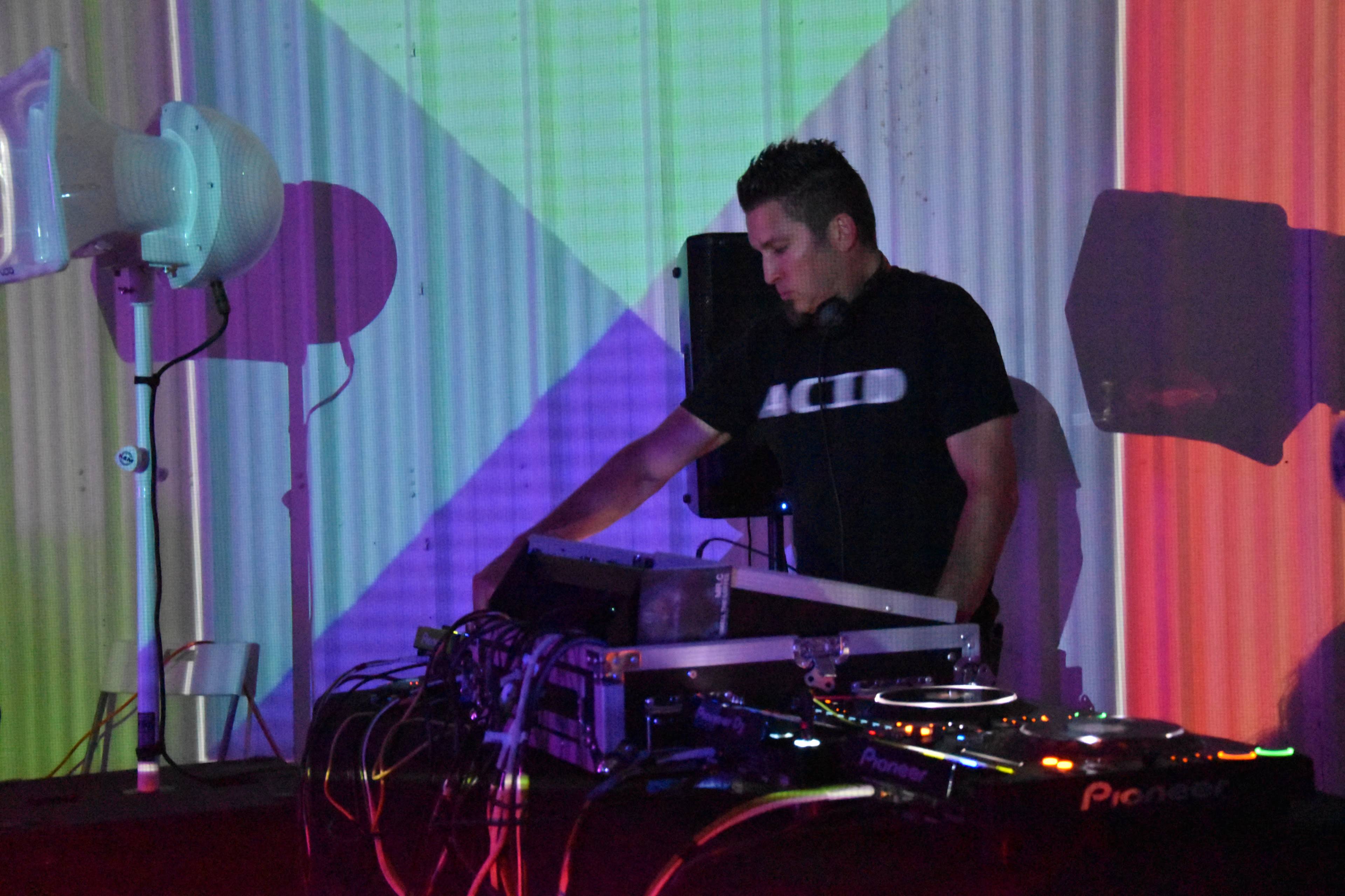 A DJ stands behind a turntable setup, manipulating equipment while colorful lights cast shadows on the wall in a dimly lit venue.