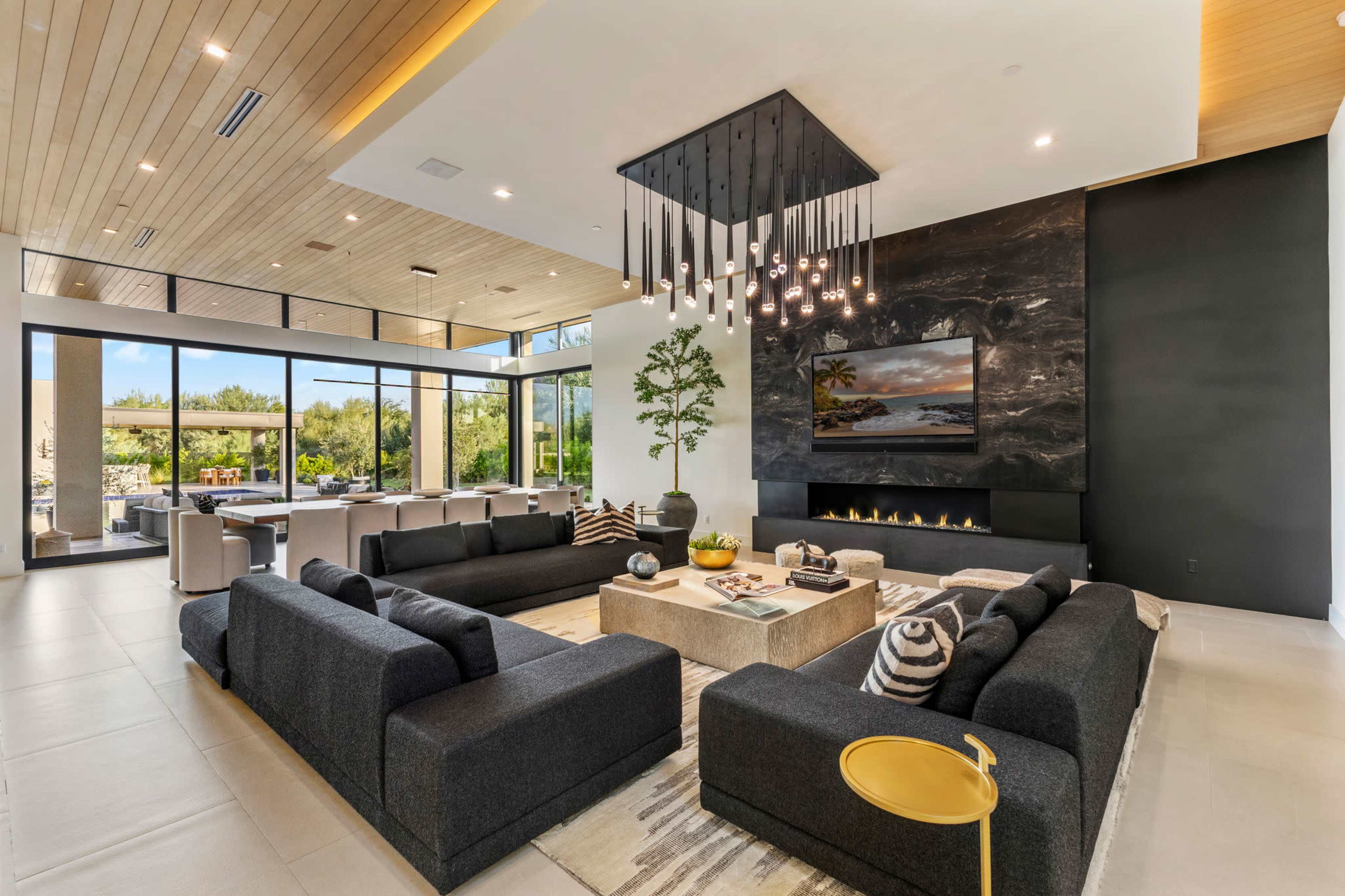 The modern living room features two black sofas facing each other around a central coffee table, with a large window showcasing an outdoor view, and a fireplace integrated into a black stone accent wall.